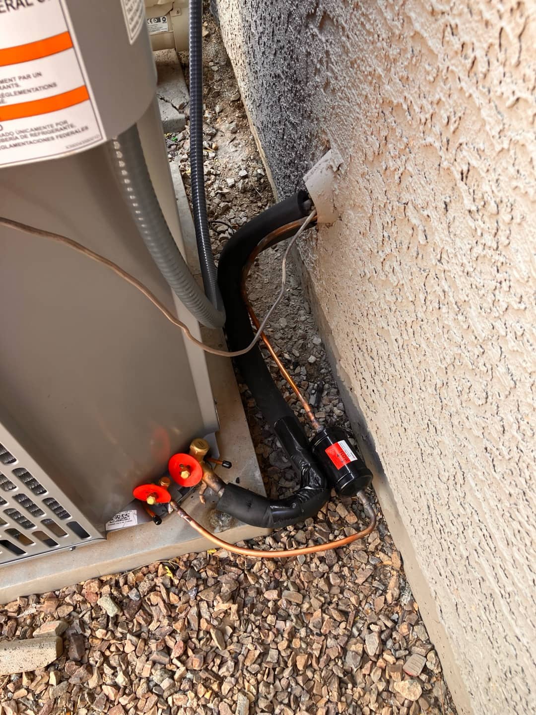 Outdoor condenser with insulated line set, suction accumulator or filter-drier, and sealed wall penetration on gravel pad.