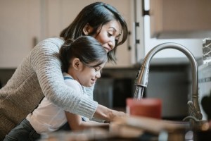 Mother helping daughter wash hands with warm water at kitchen sink, suggesting hot water heater replacement.