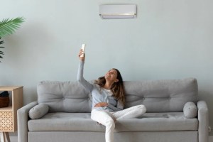 Woman sitting on a couch using remote to operate wall-mounted heating system.