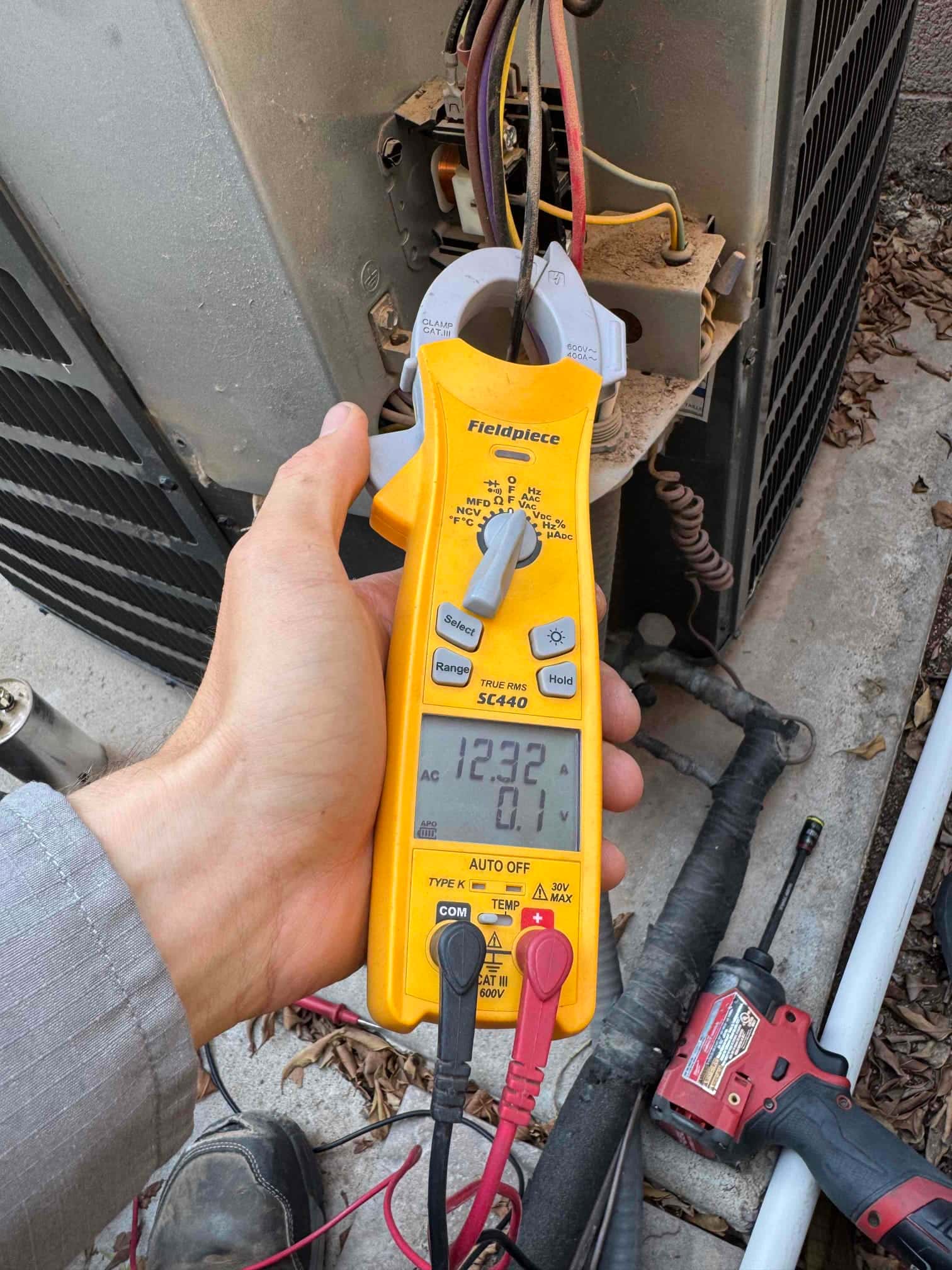 Clamp meter displays 12.32 A on a compressor lead at the outdoor condenser with test leads connected.