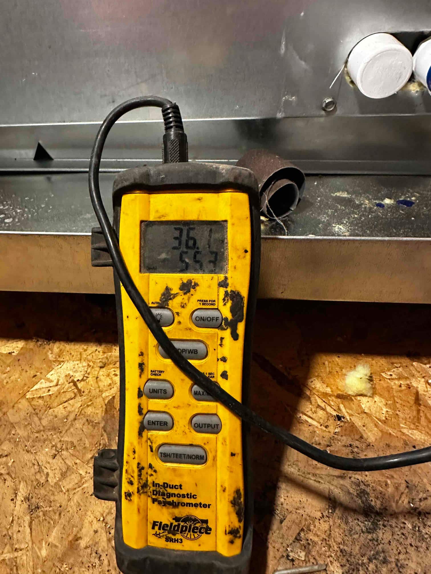Digital thermometer-hygrometer measuring air at the duct, display shows 36.1 and 55.7 on separate lines during airflow testing.