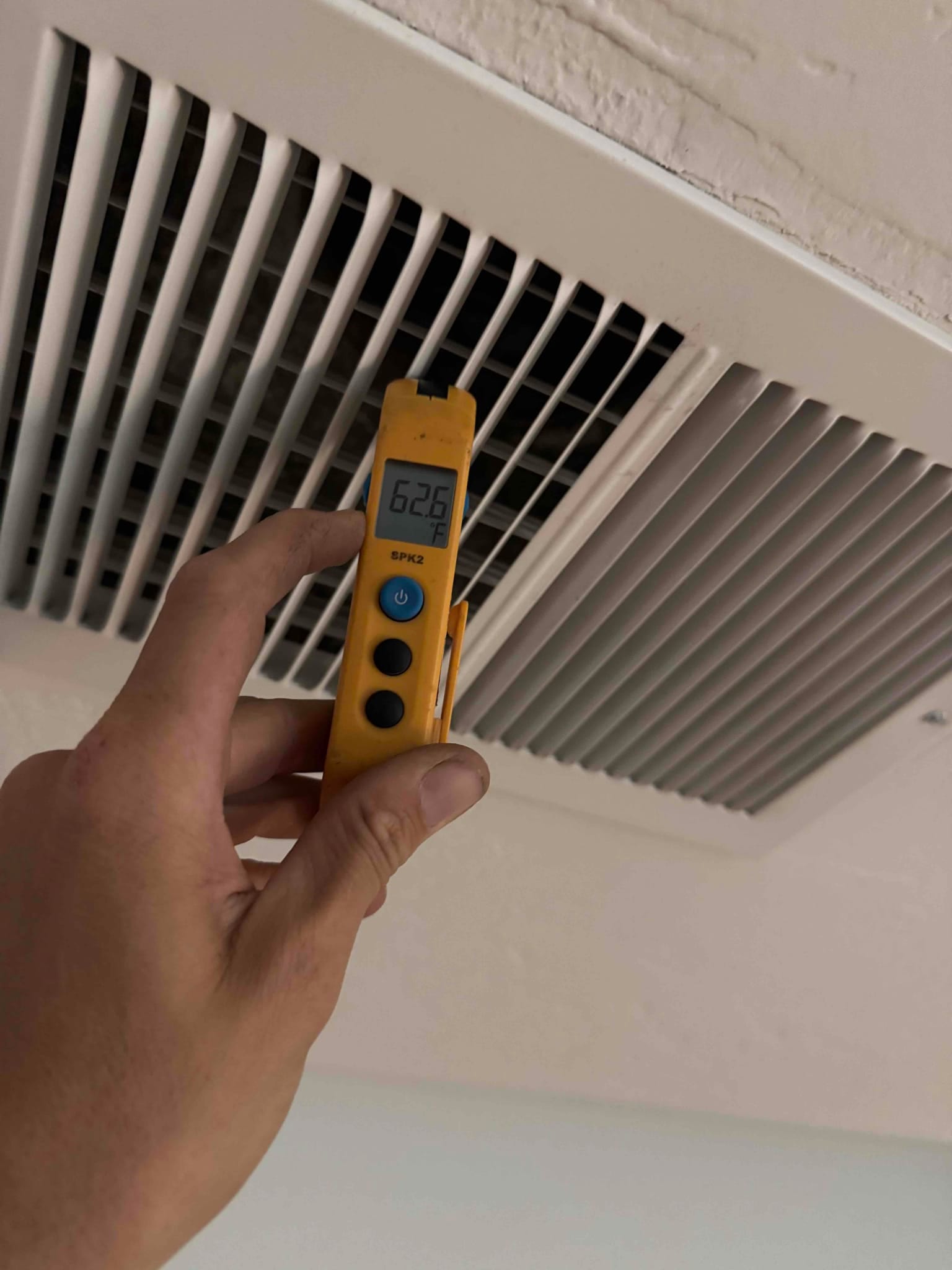 Digital thermometer shows 62.6 °F at the ceiling supply vent, measuring the temperature of the discharged air.