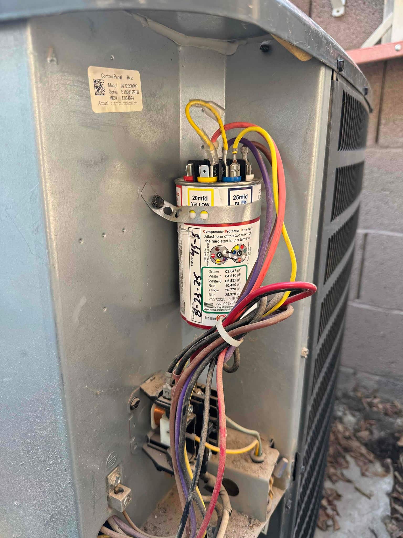 Dual-run capacitor installed in outdoor condenser, wiring connected to labeled terminals; printed rating shows 20/25 μF with wiring diagram.