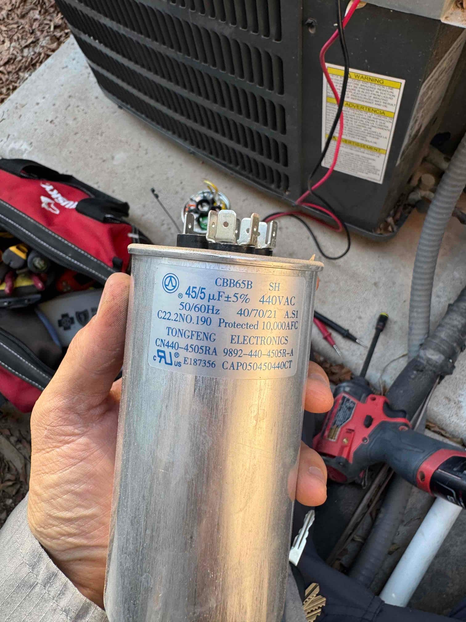 Technician holds a dual run capacitor rated 45/5 μF, 40/70/21, 40VAC, showing three terminals and printed specifications.