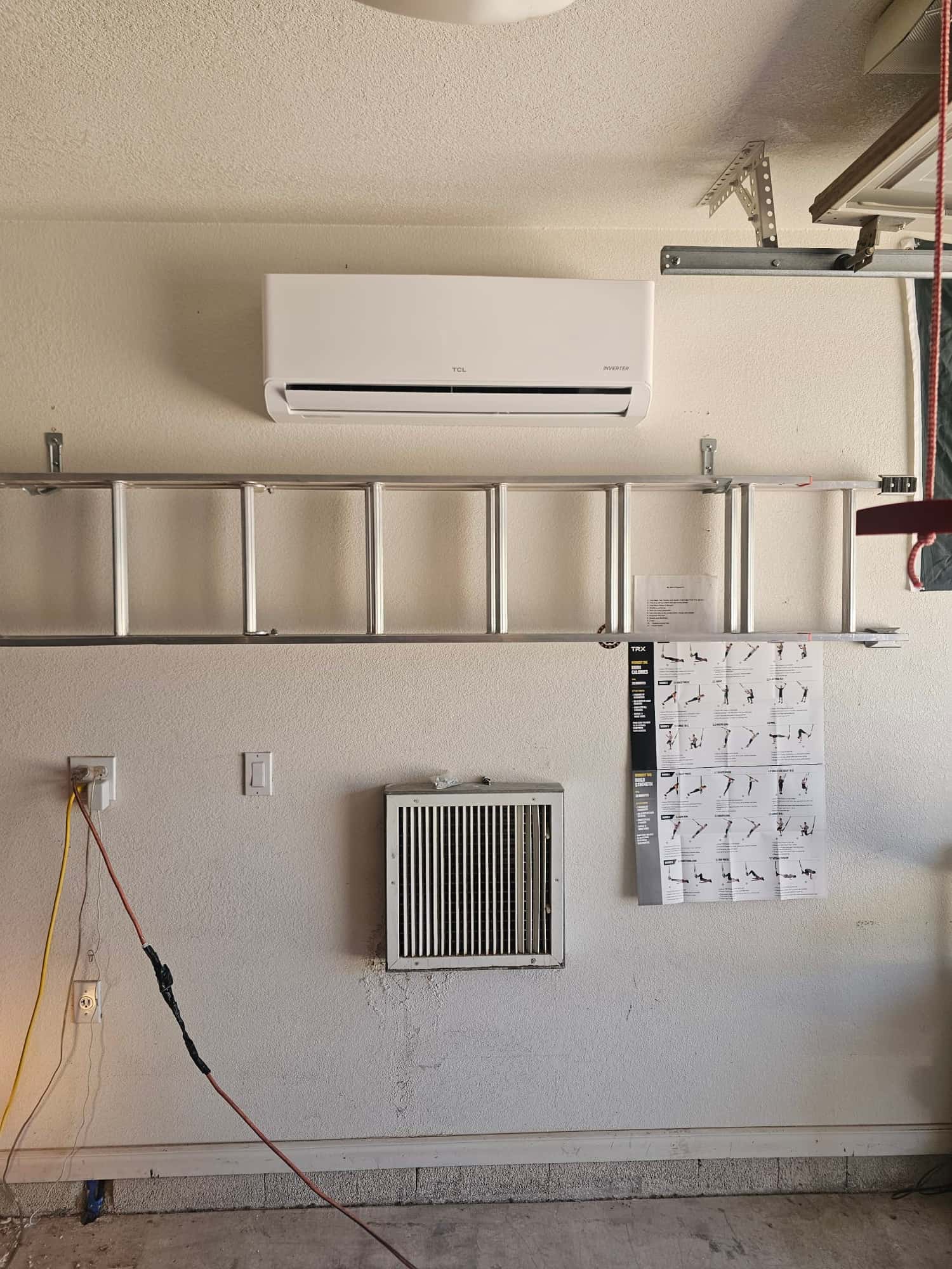 Wall-mounted mini-split indoor unit installed in a garage, with mounting hardware and ladder below, showing clean, finished placement.