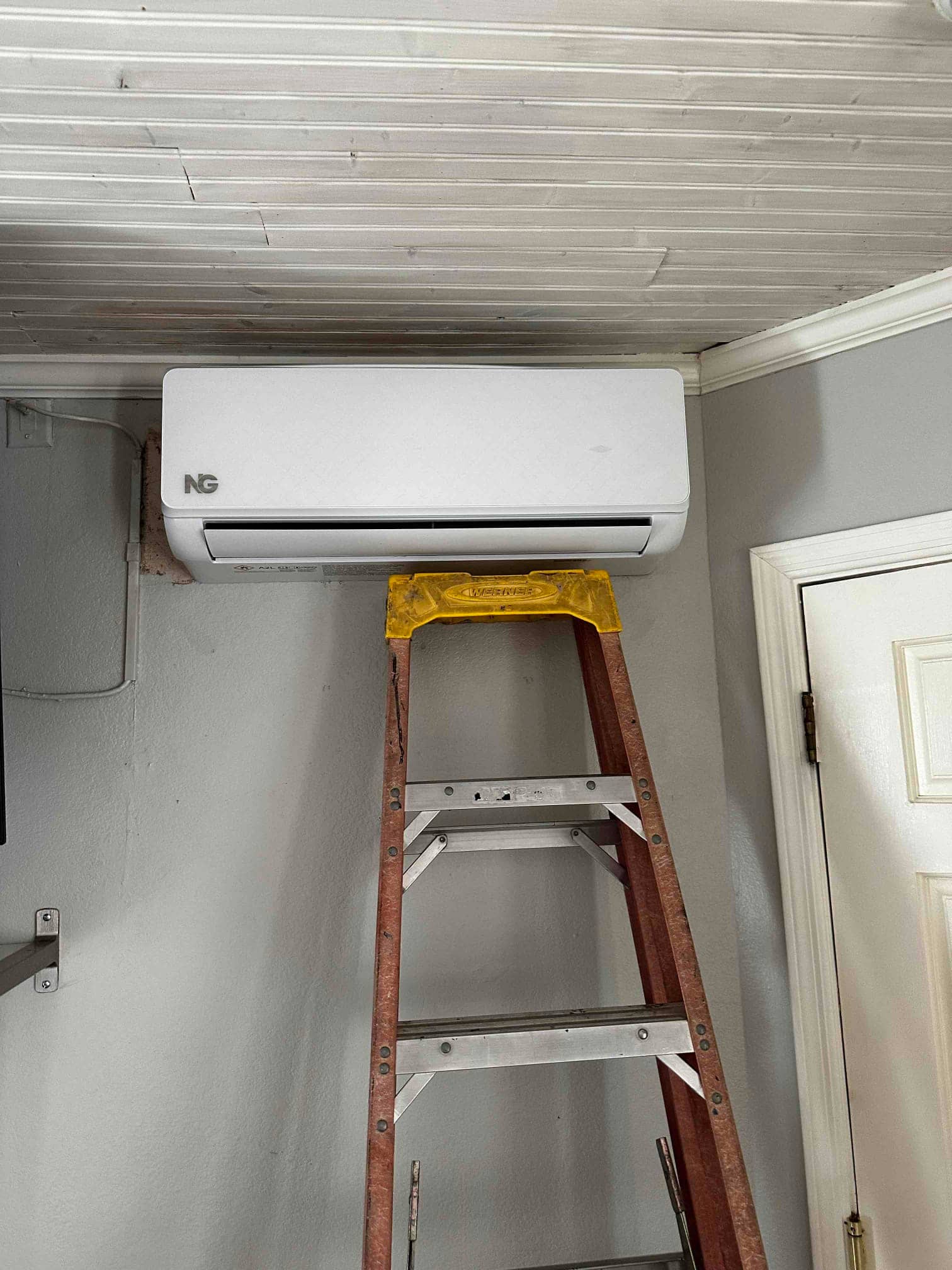 Wall-mounted mini-split indoor unit newly installed near the ceiling, with a ladder positioned beneath it for final adjustments.