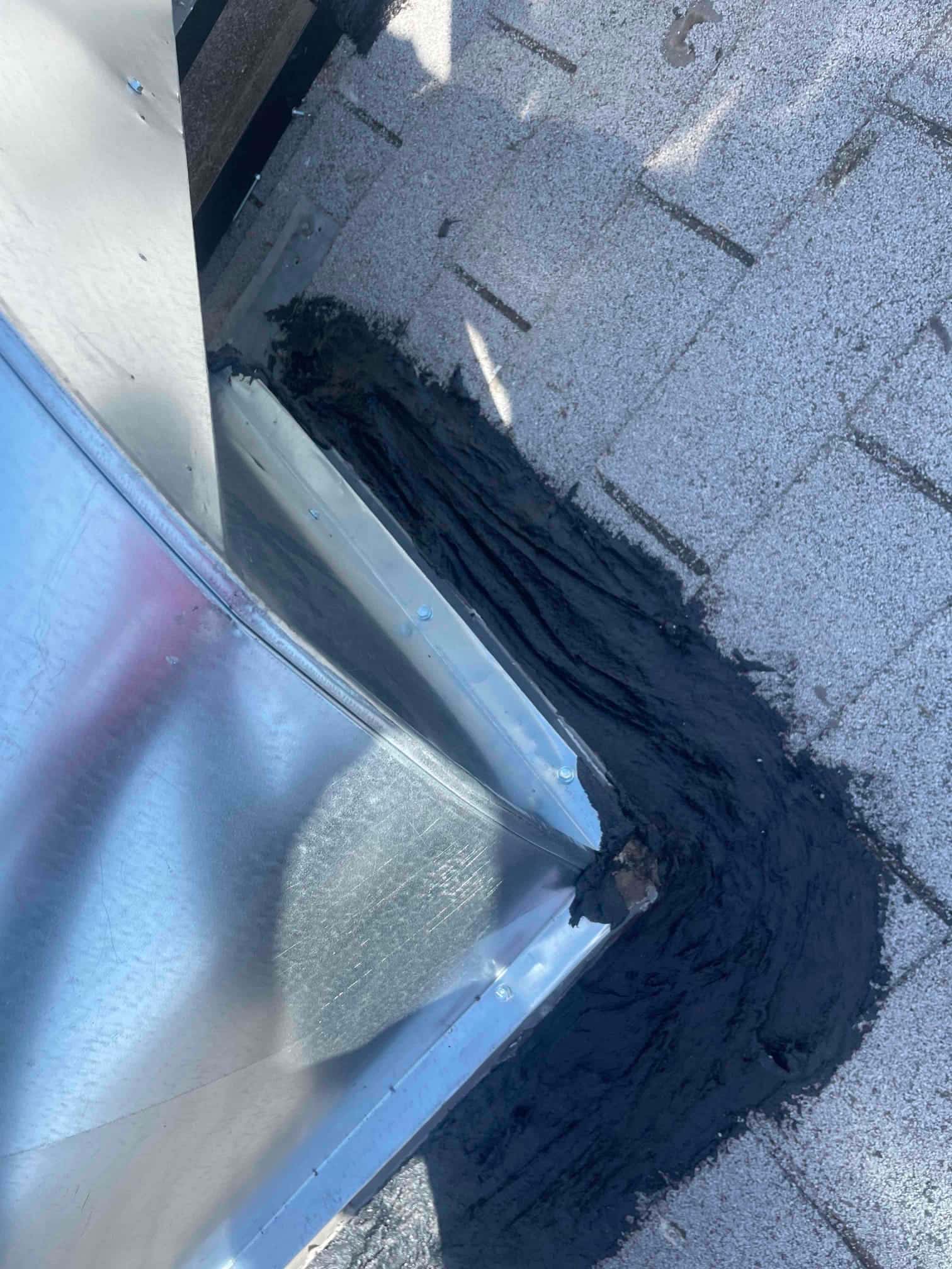 Sheet metal duct curb flashing on a shingle roof, sealed with roofing mastic around the base to prevent water intrusion.