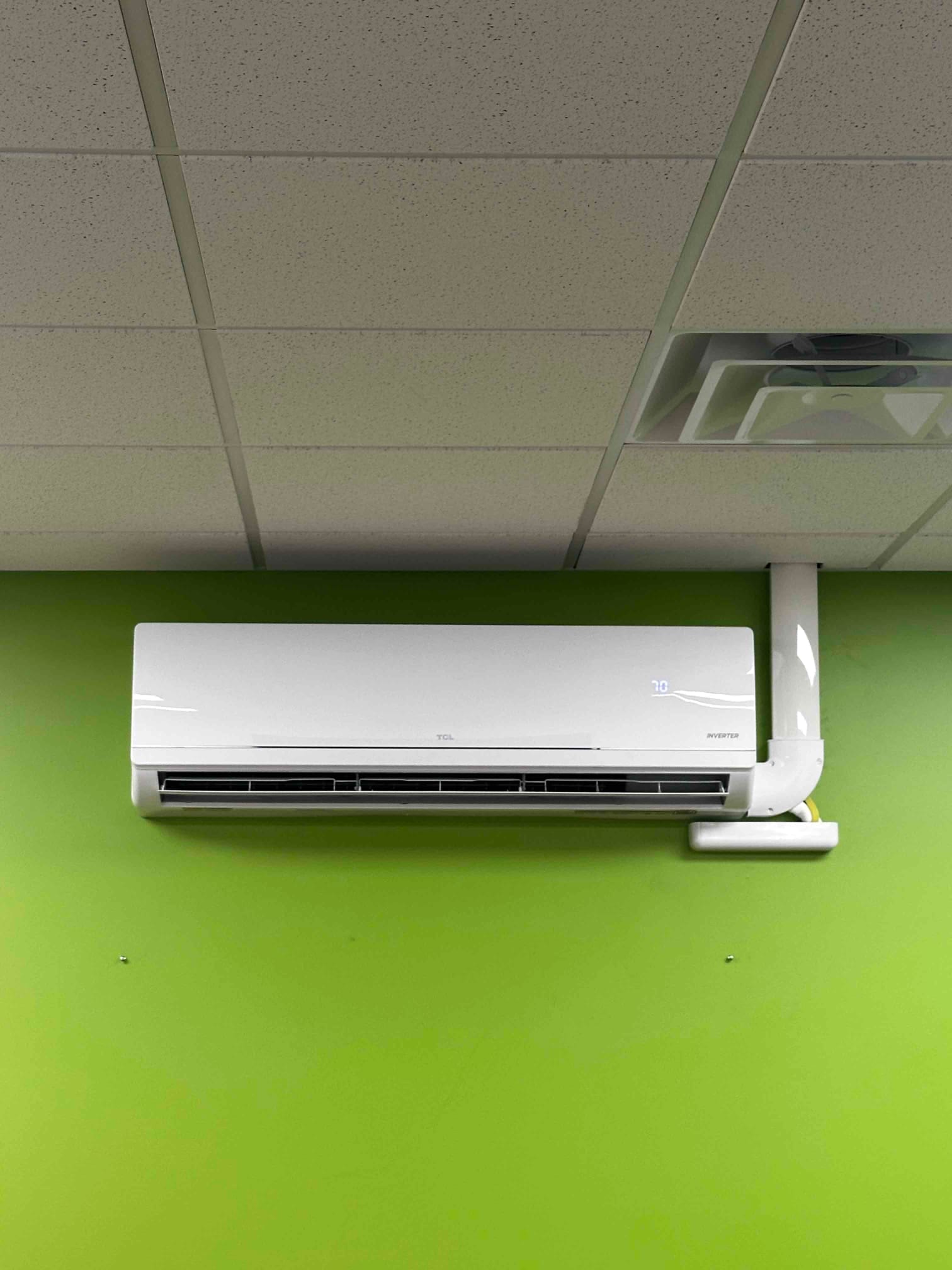 Wall-mounted mini-split indoor unit installed on a green interior wall with line-set cover and drain exiting to the right.