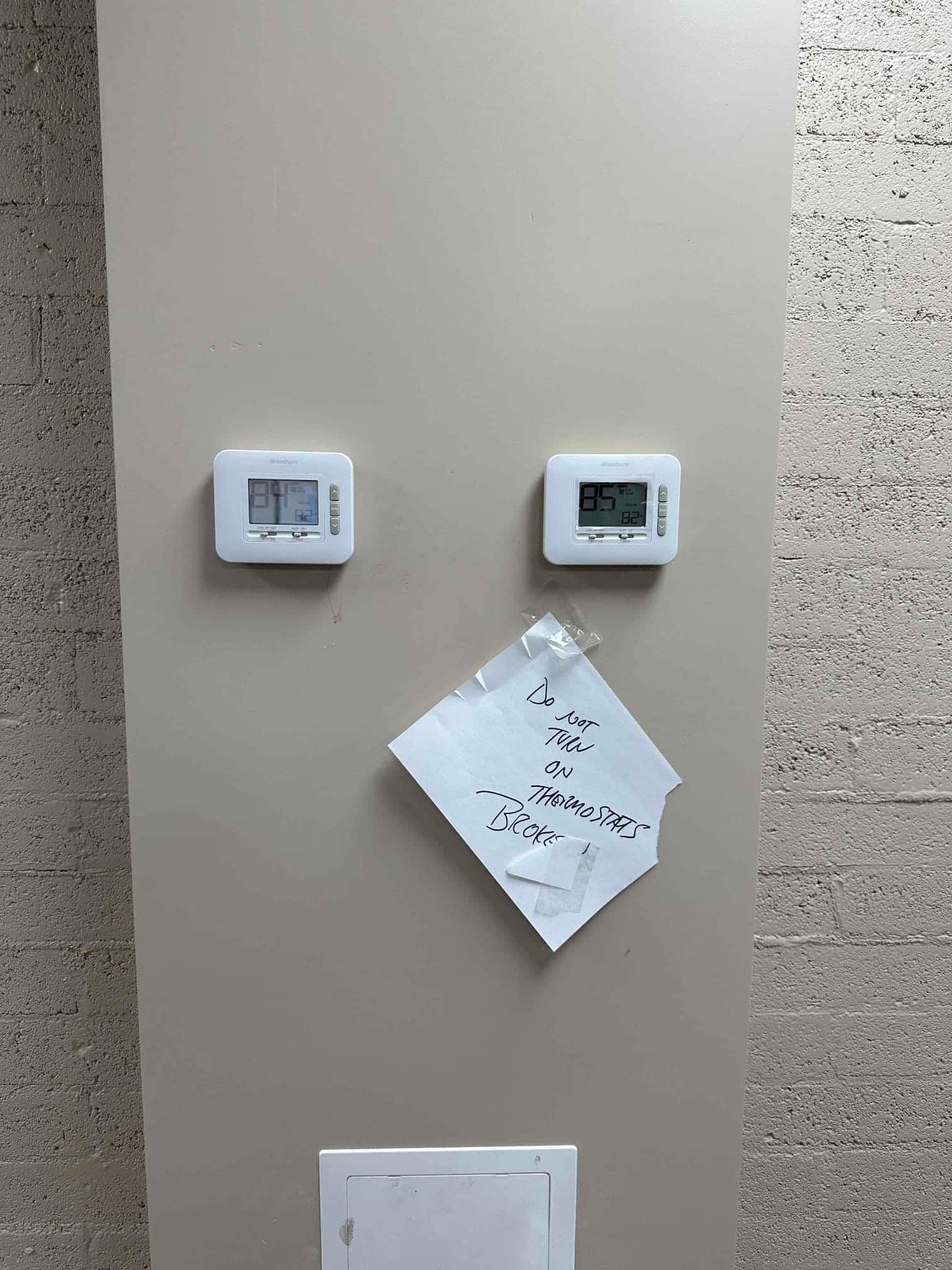 Two wall-mounted thermostats installed side by side with a handwritten note taped nearby instructing not to turn them on.
