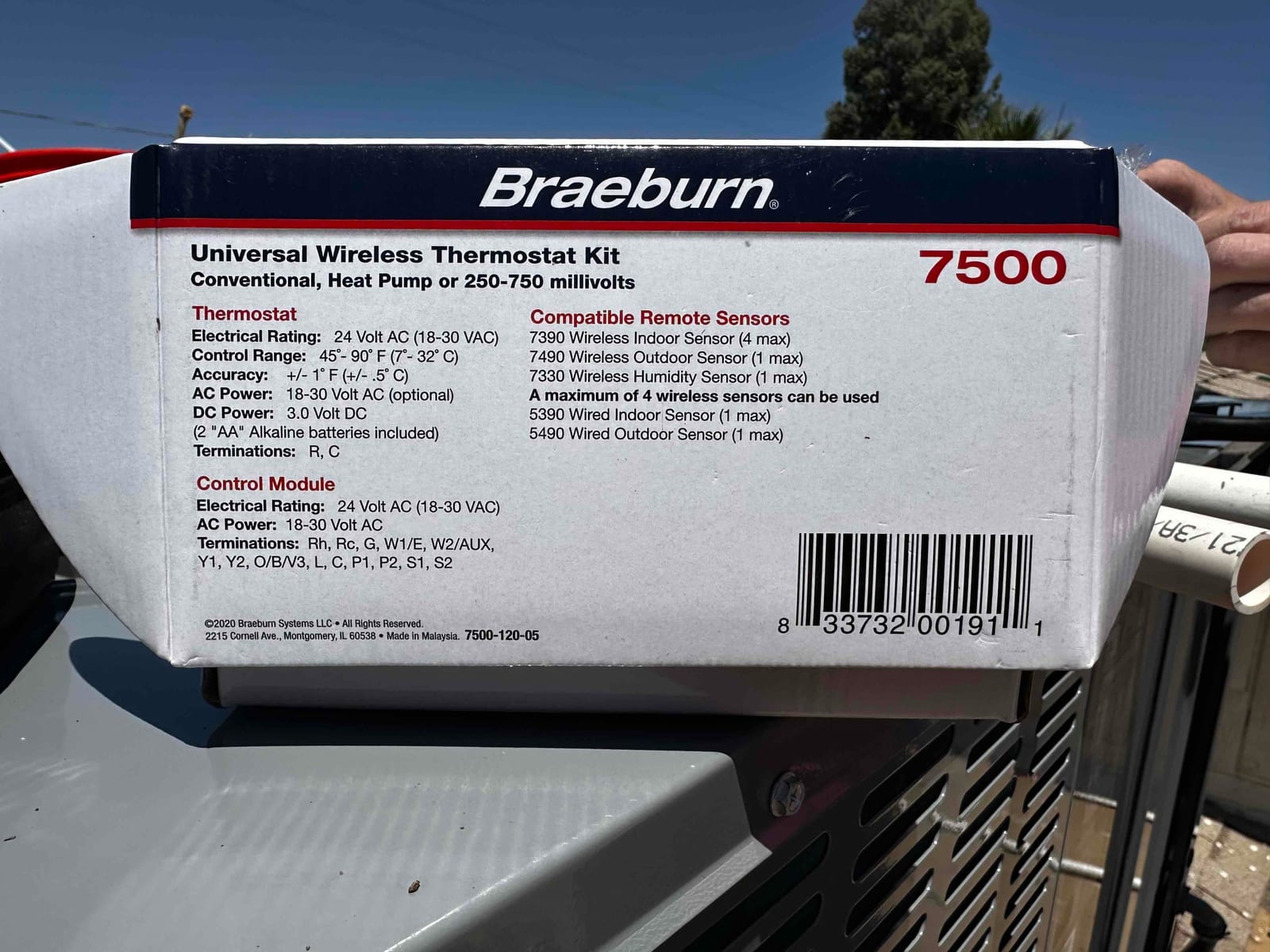 Box for a universal wireless thermostat kit resting on top of outdoor HVAC equipment during installation.