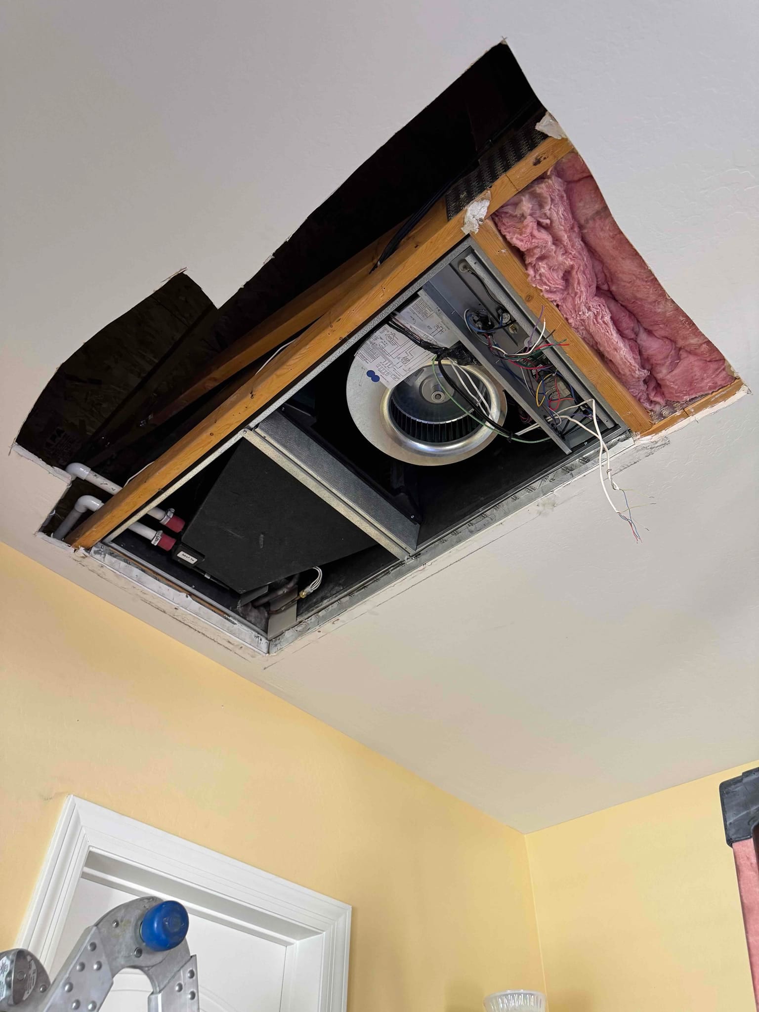 Ceiling-mounted air handler installed in an attic opening with visible blower, insulation, drain line, and wiring awaiting final paneling.