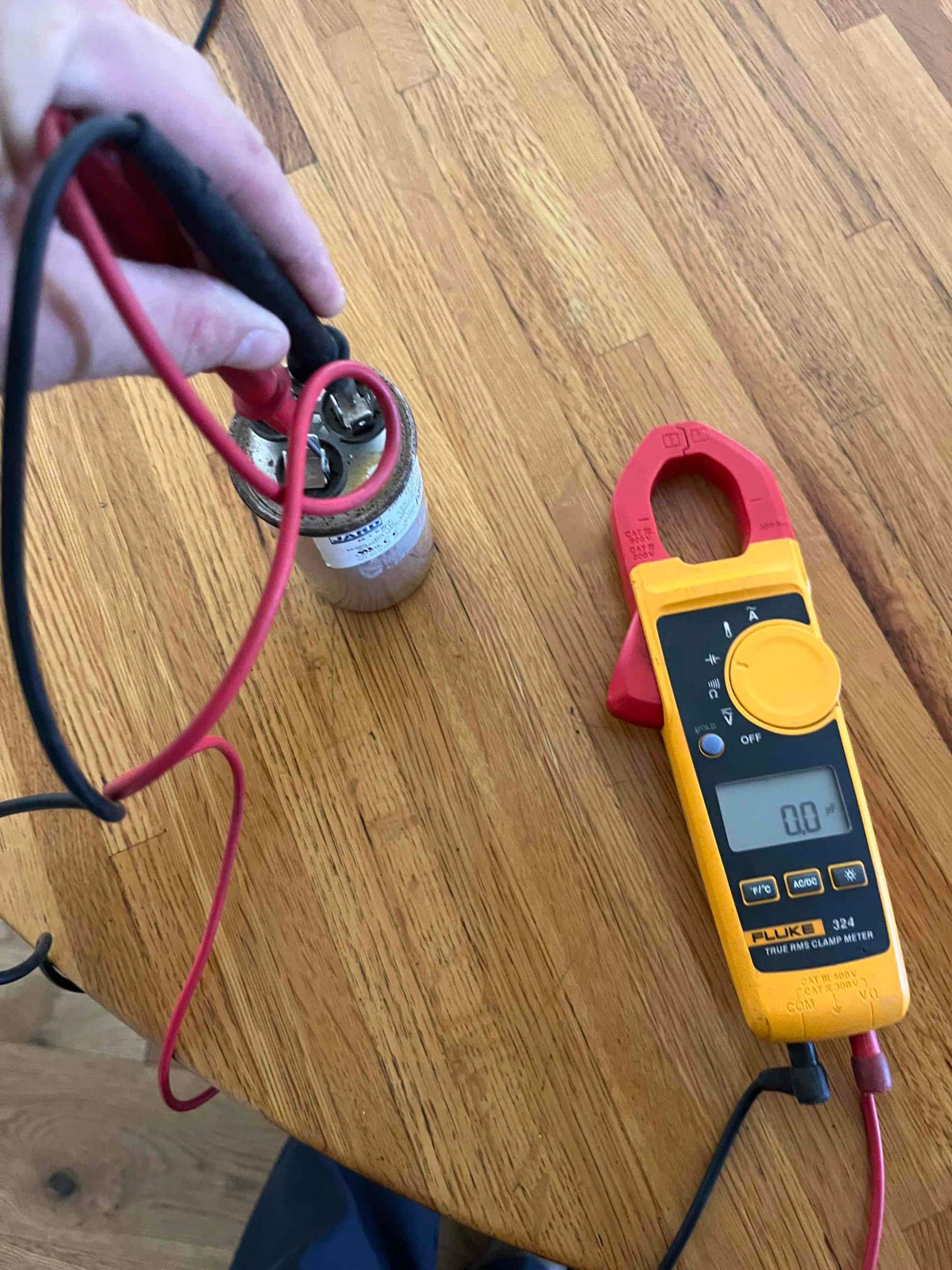 Clamp meter display shows 0.0 Ω while test leads contact terminals on a dual-run capacitor.