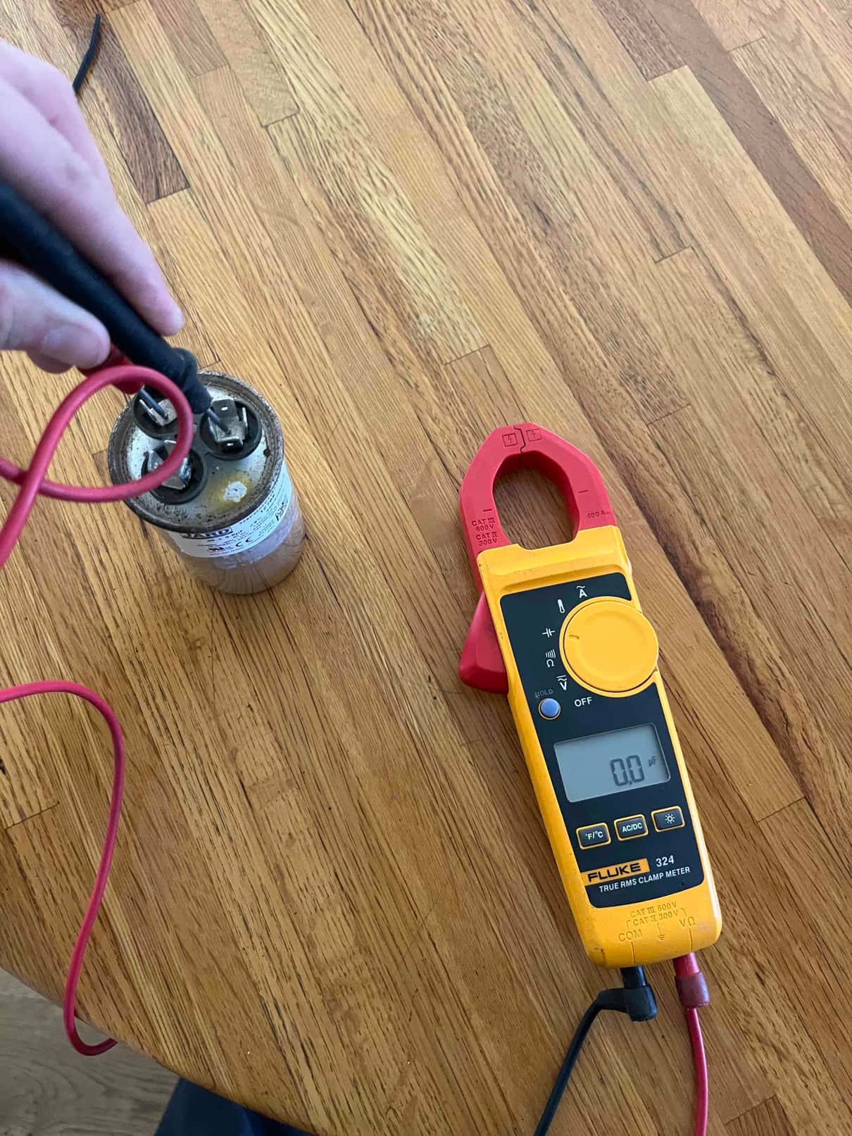 Clamp meter shows 0.1 A while test leads contact terminals on a removed dual-run capacitor.