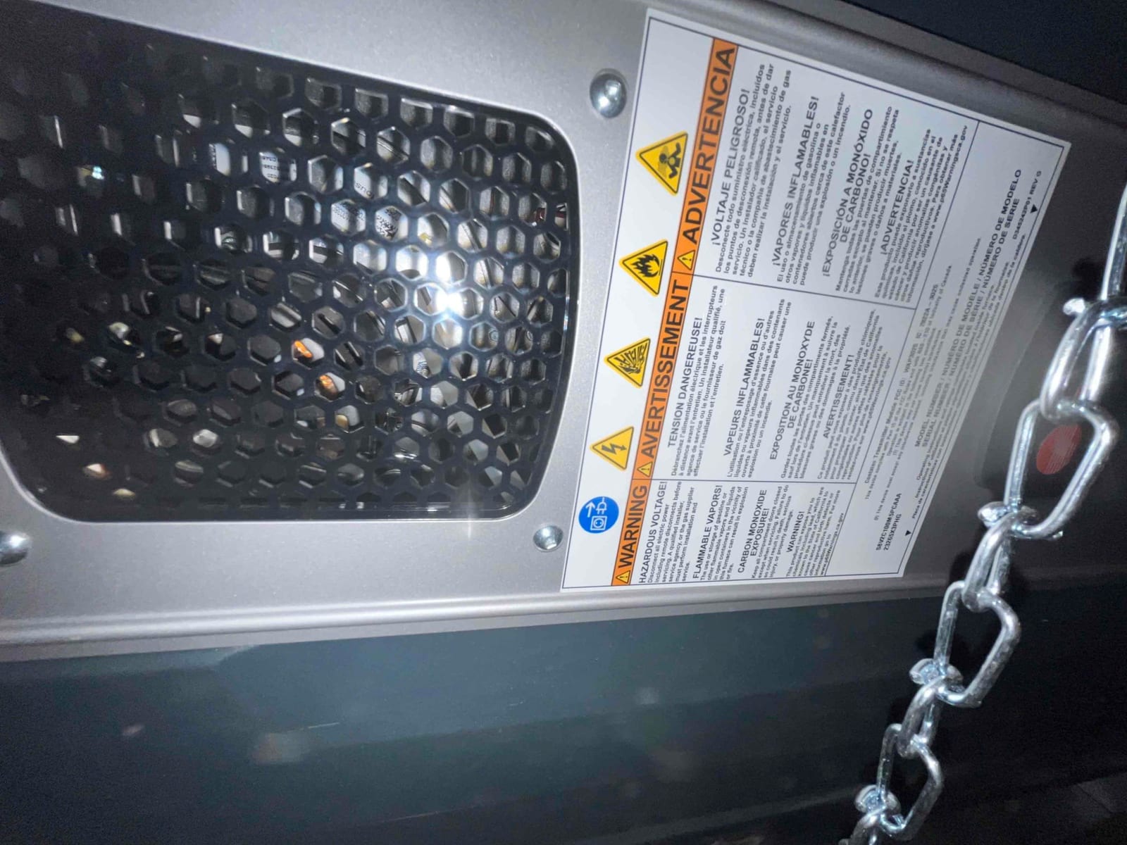 Close-up of an HVAC unit’s access panel with ventilation grille and multilingual safety warning label beside a hanging chain.