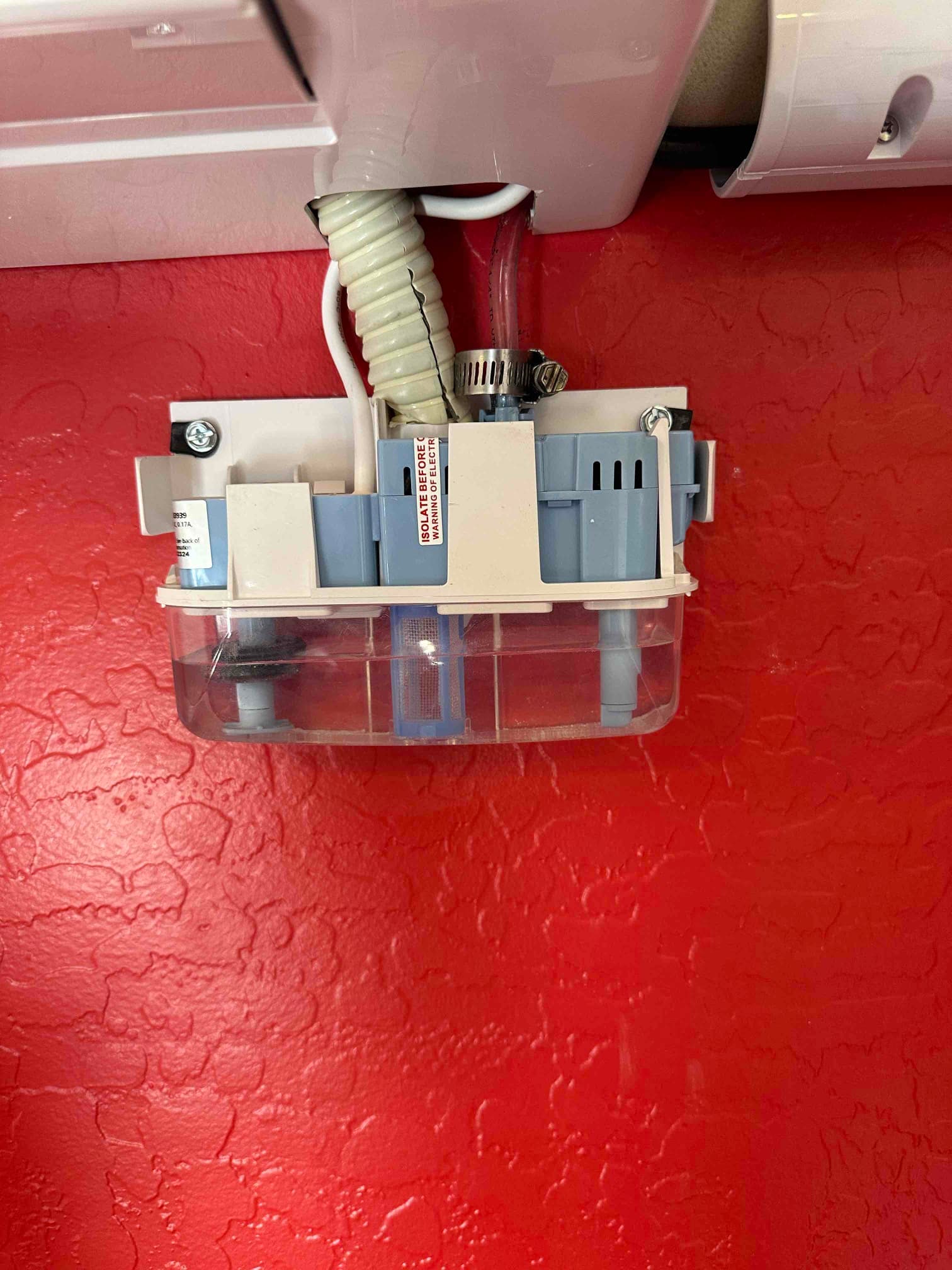 Condensate pump mounted beneath a wall-mounted indoor unit, with drain tubing, wiring, and clamp connections visible against a red wall.