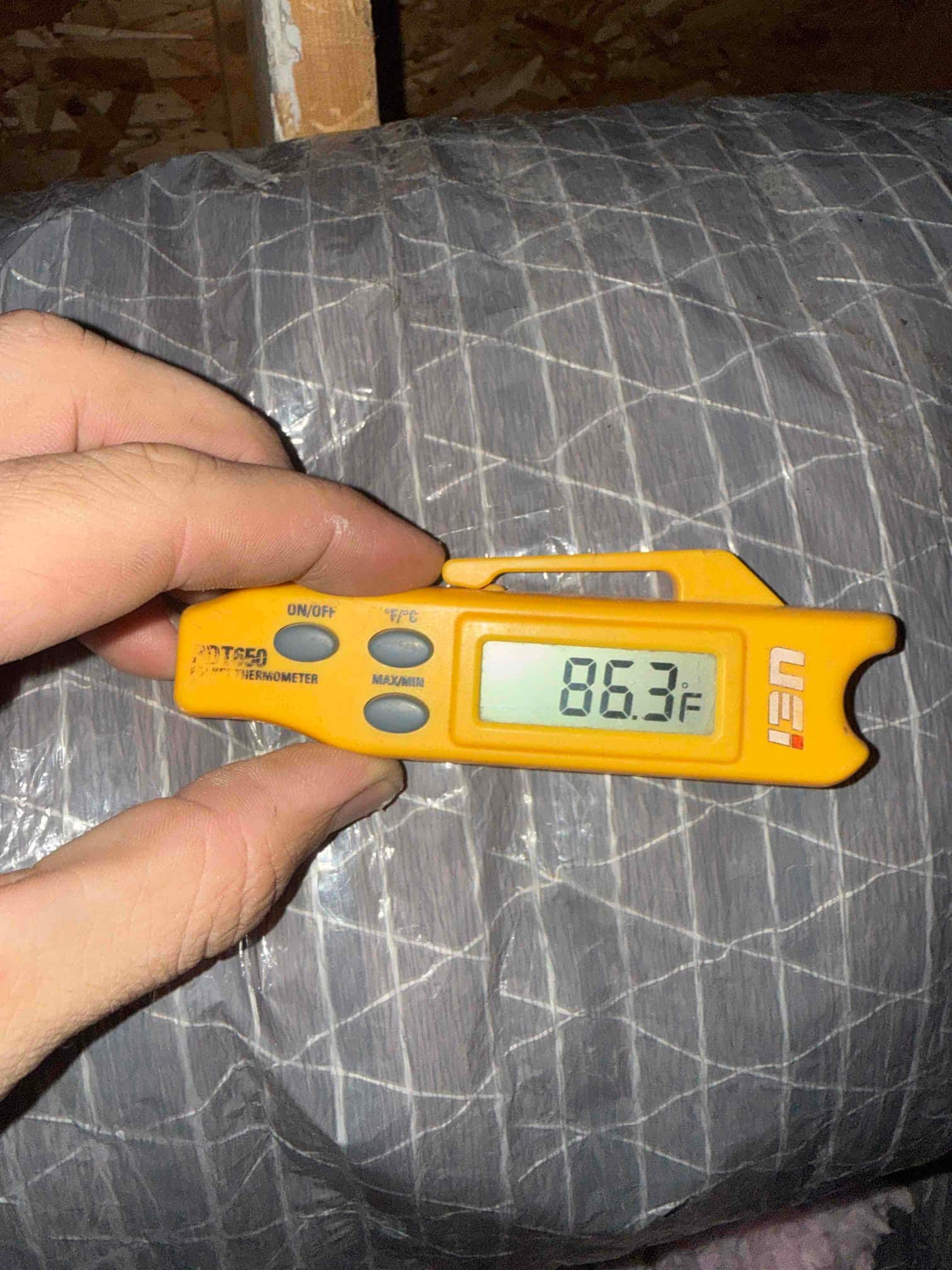 Digital thermometer shows 86.3°F while checking air temperature at insulated flexible duct in the attic.