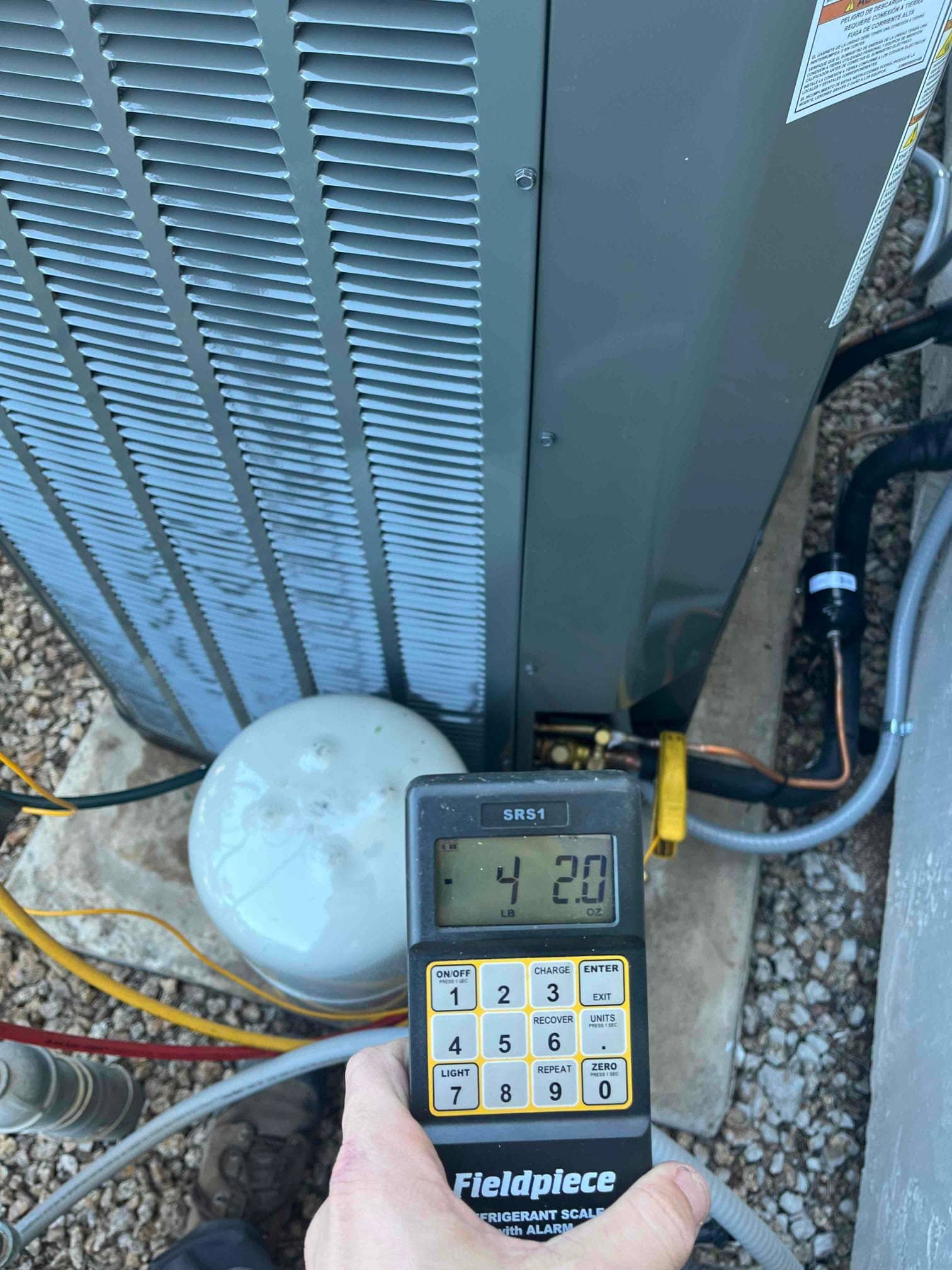 Handheld refrigerant scale displays 4 lb 20 oz near the outdoor condenser during system charging at the installation site.
