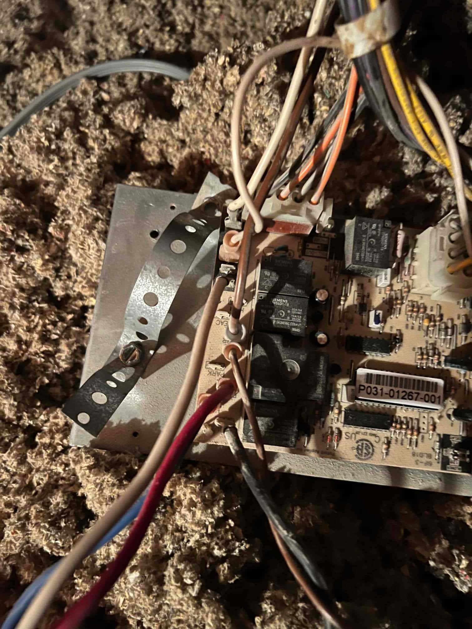 HVAC control board with multiple relays and wire connections exposed beside attic insulation and a perforated metal mounting strap.