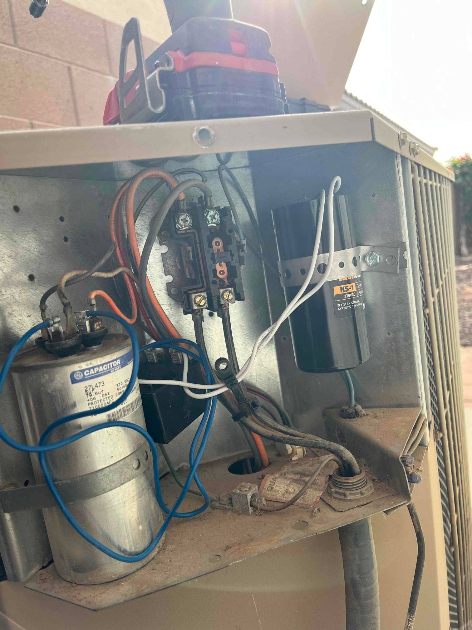 Open condenser control panel showing contactor, dual-run capacitor, and start capacitor with connected wiring and dust accumulation.