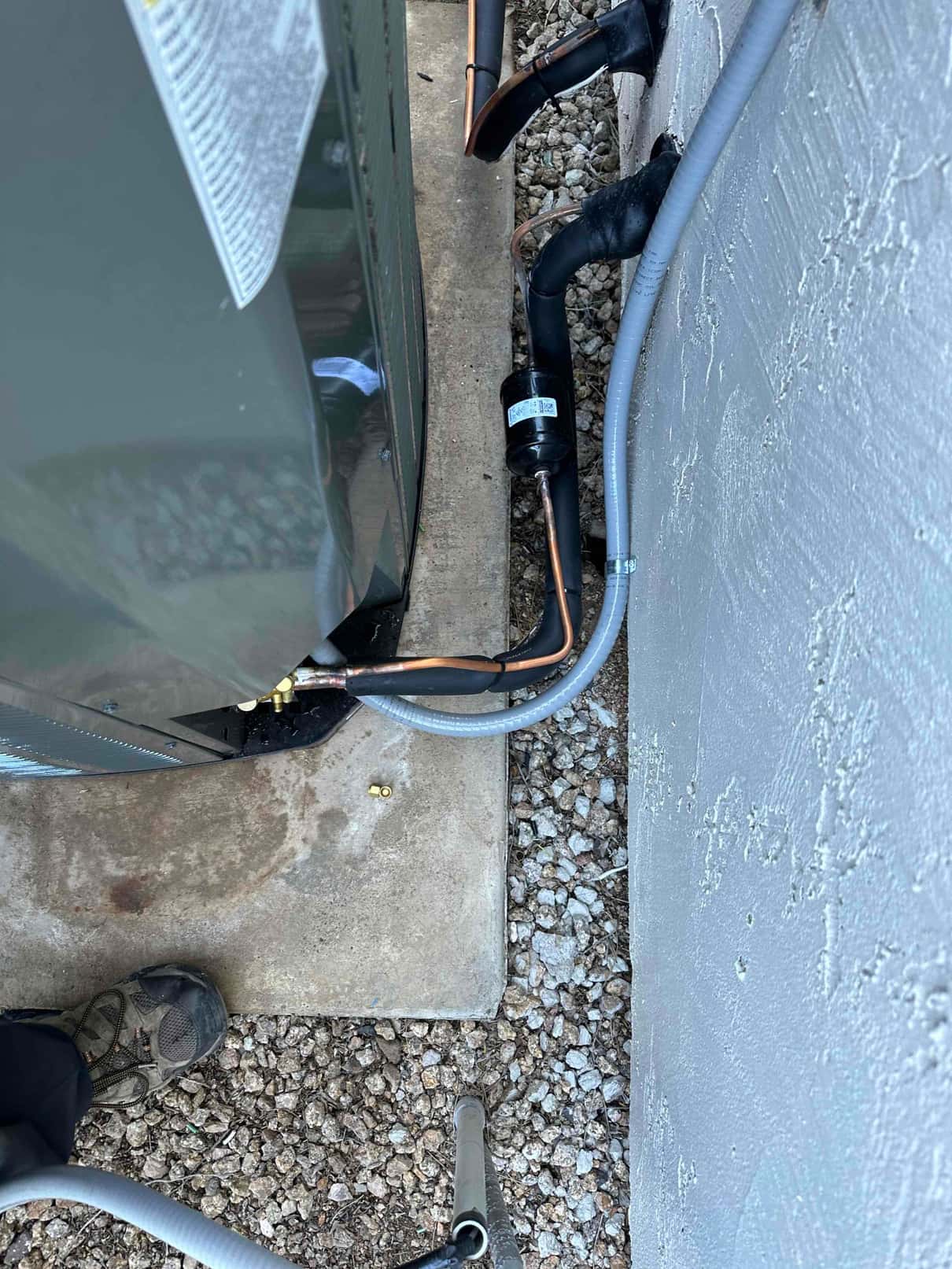 Outdoor condenser set on a concrete pad with insulated refrigerant lines, filter-drier, and electrical conduit entering the wall.