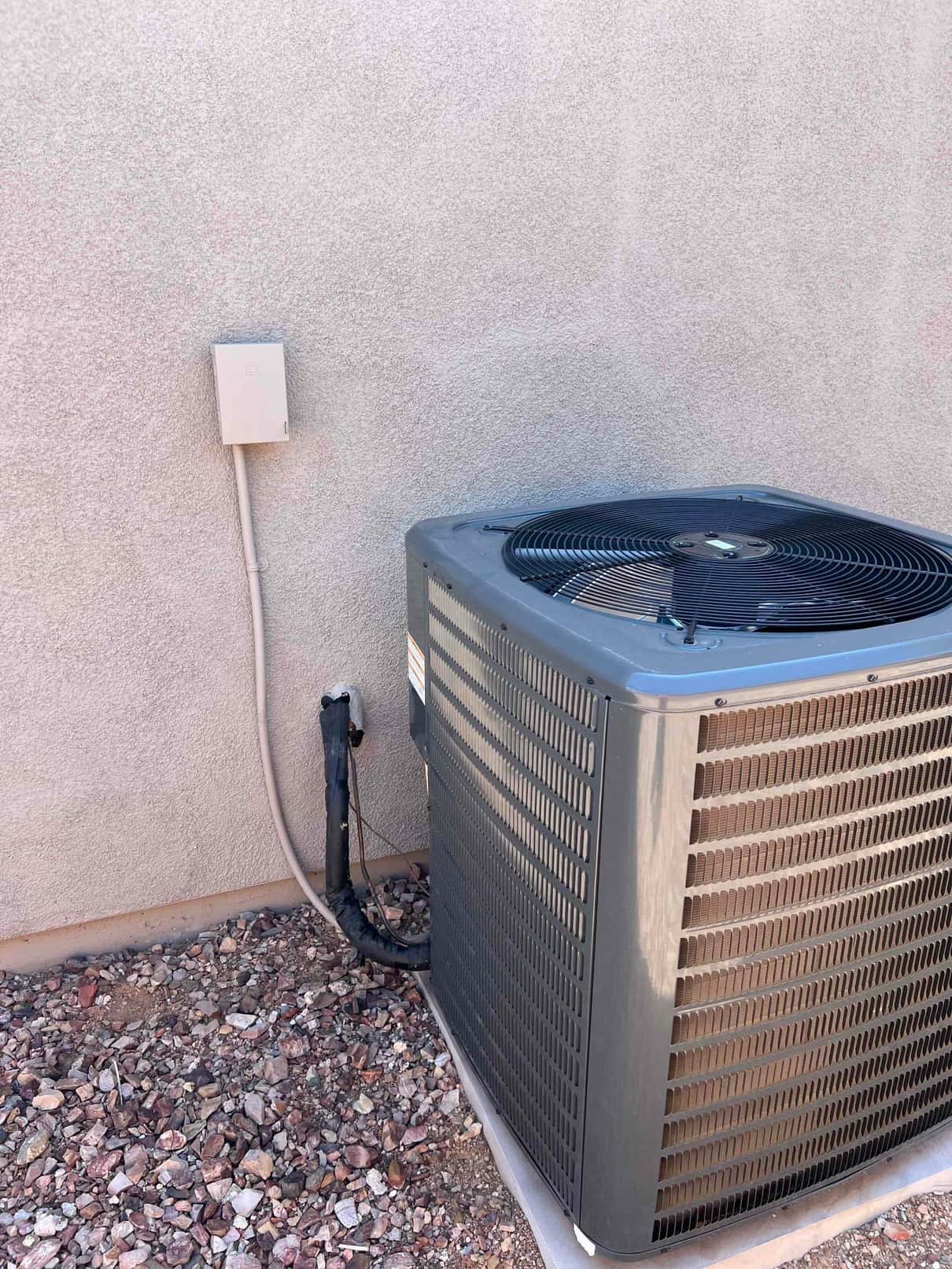 Outdoor condenser unit beside wall with disconnect box, insulated refrigerant lines, and protective top fan grille.