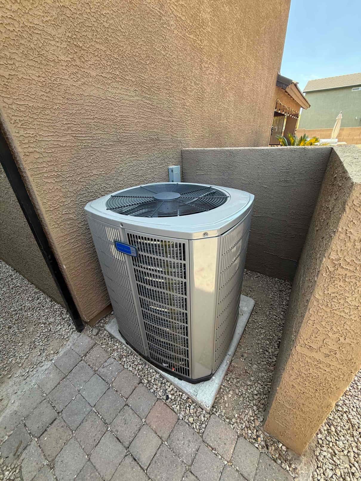 Outdoor heat pump condenser set on a level pad within a side yard alcove, with protective wall and paver walkway.