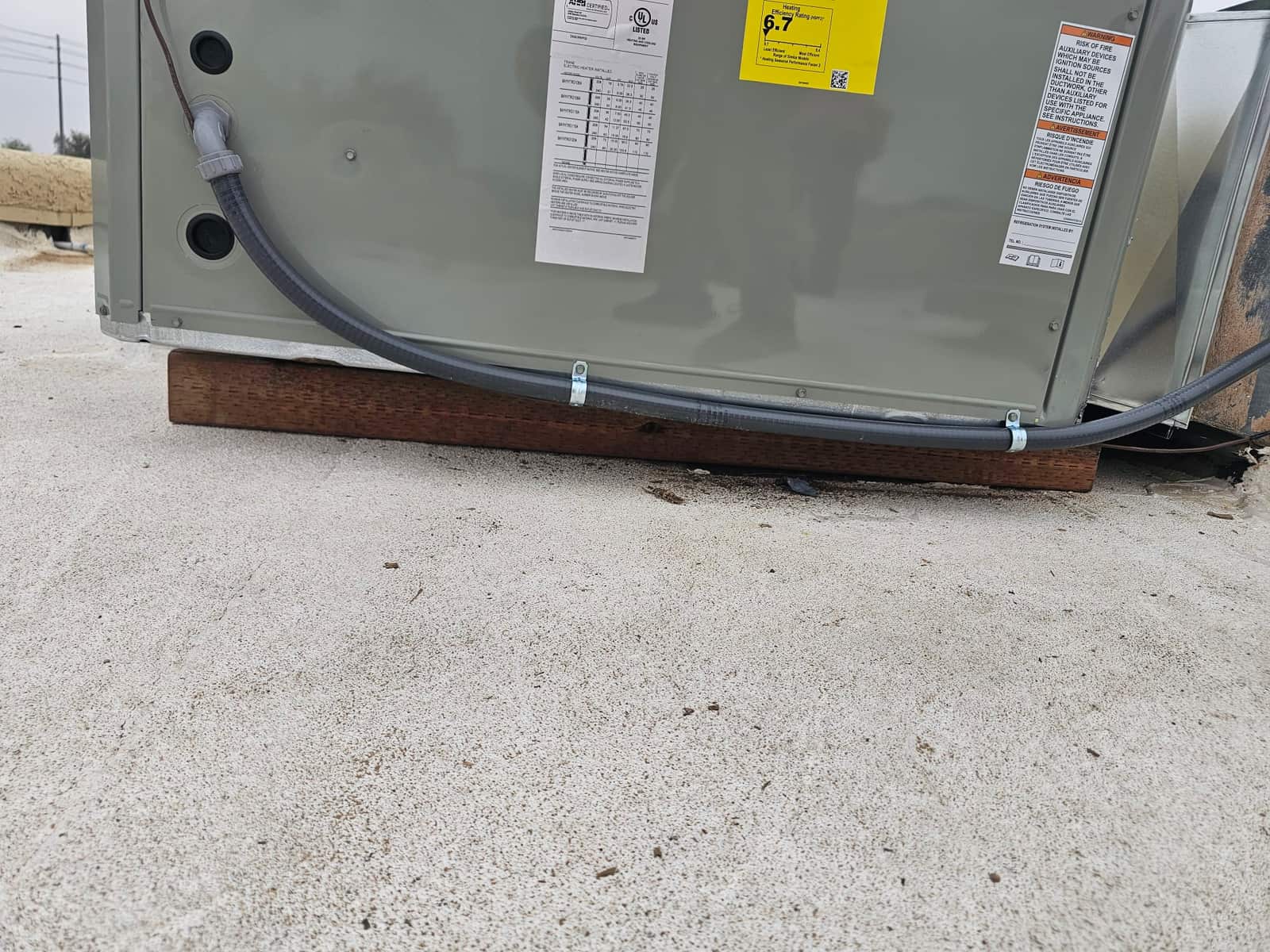 Outdoor HVAC unit base set on wood risers with flexible electrical conduit secured to the cabinet on a rooftop surface.