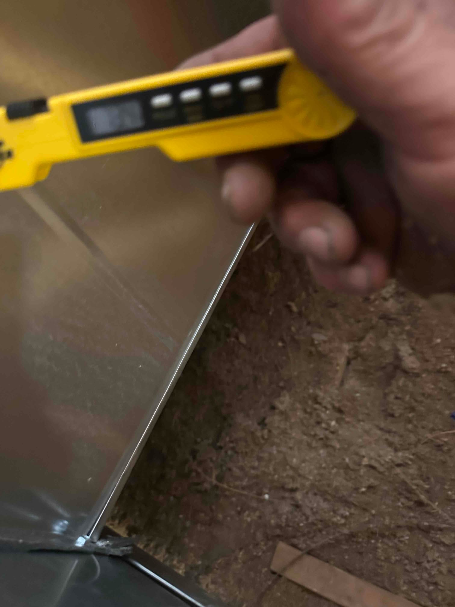 Sheet metal duct transition is positioned over the air handler with a digital thermometer held nearby during installation.