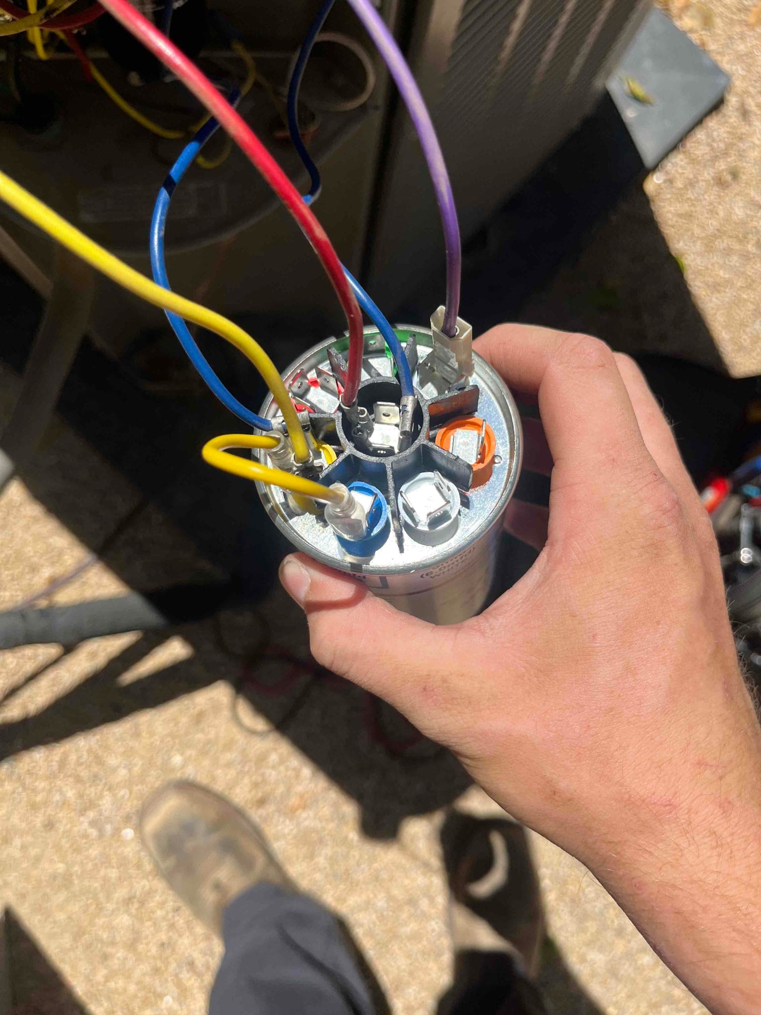 Technician holds a dual-run capacitor with connected color-coded wires near the outdoor condenser unit during repair.