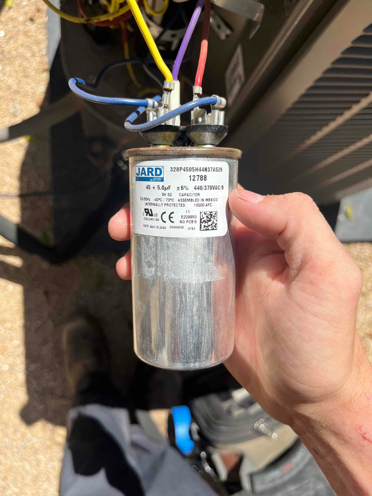 Technician holds a dual run capacitor labeled 45/5 μF at 440VAC with connected compressor and fan wires.