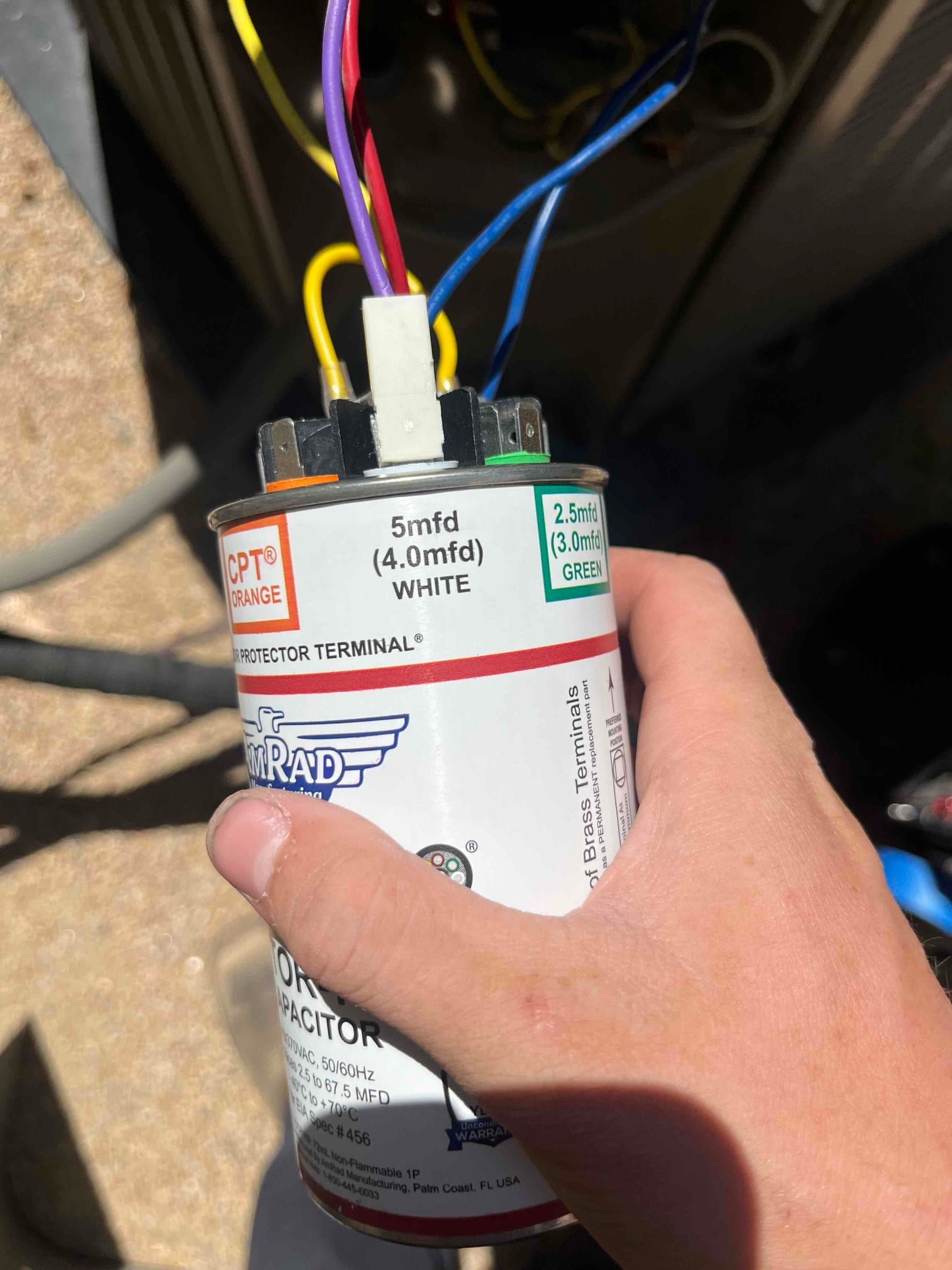 Technician holds a dual-run capacitor showing printed ratings: 5 mfd (4.0 mfd) and 2.5 mfd (3.0 mfd) with color-coded terminals.