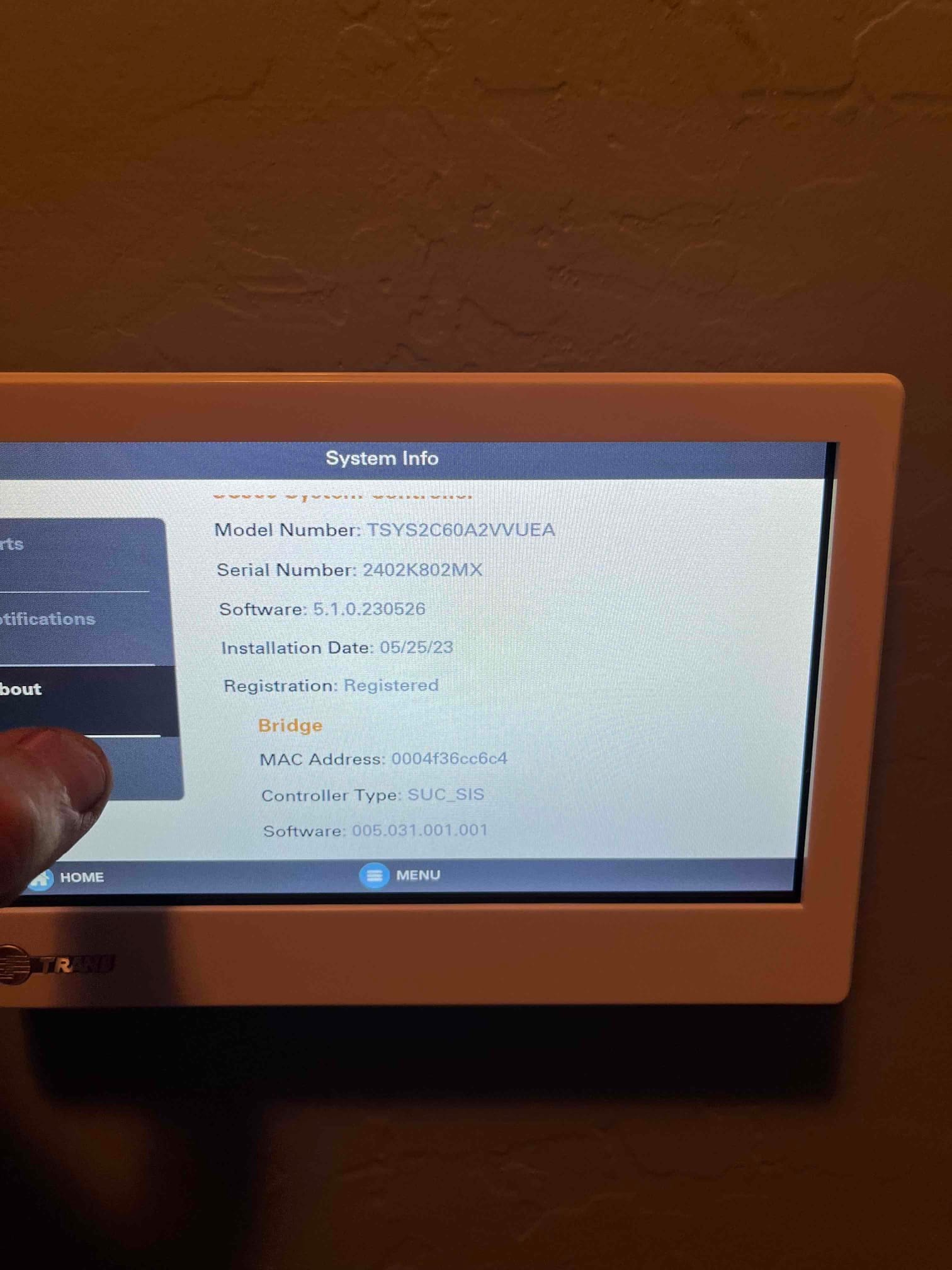 Touchscreen thermostat displays system information and registration status after installation, with a technician’s finger selecting the menu.