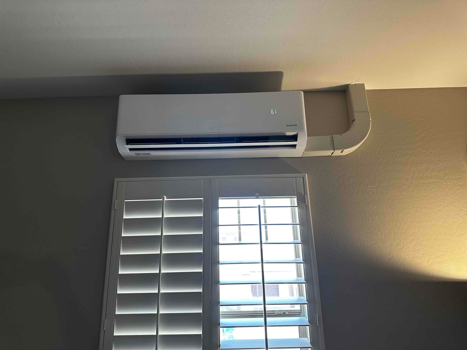 Wall-mounted mini-split indoor unit installed above a window with routed line-set cover and louvered supply outlet.