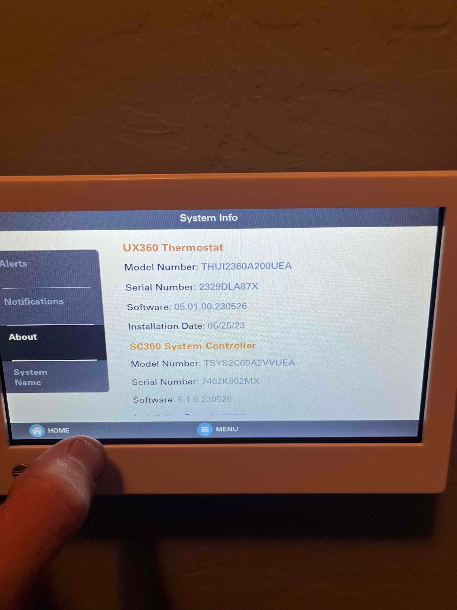 Wall-mounted touchscreen thermostat displays system information menu, showing model, serial, and software details for the controller.