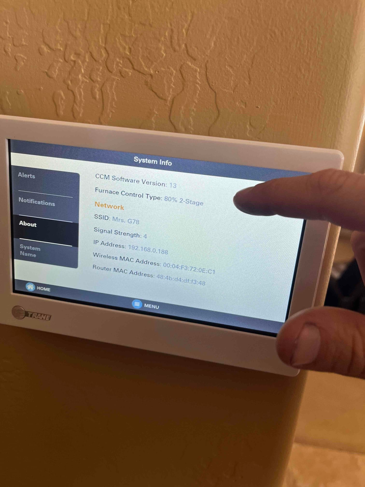 Wall-mounted touchscreen thermostat displays system information and network details during setup, with a technician’s hand interacting with the menu.