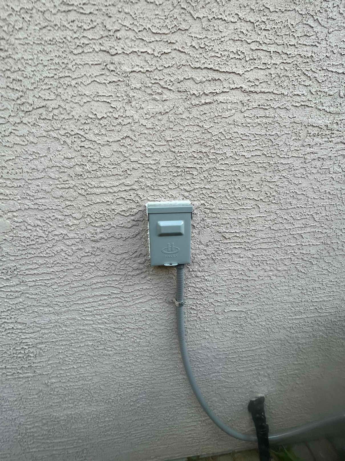 Weatherproof HVAC disconnect box mounted on exterior stucco wall with flexible conduit routed to the condenser location.