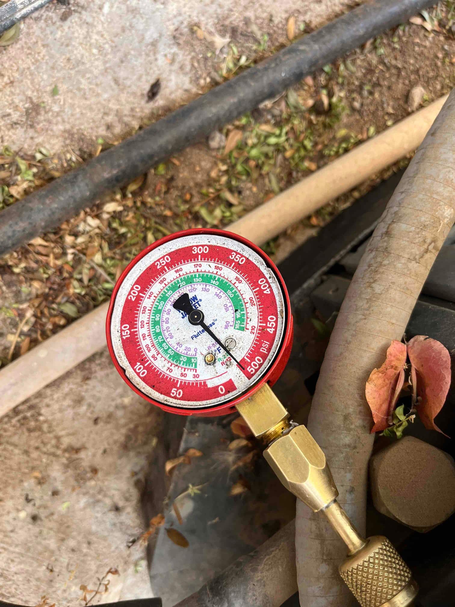 Analog refrigerant manifold gauge connected to an HVAC service port, showing pressure scales with colored ranges and needle indicator.
