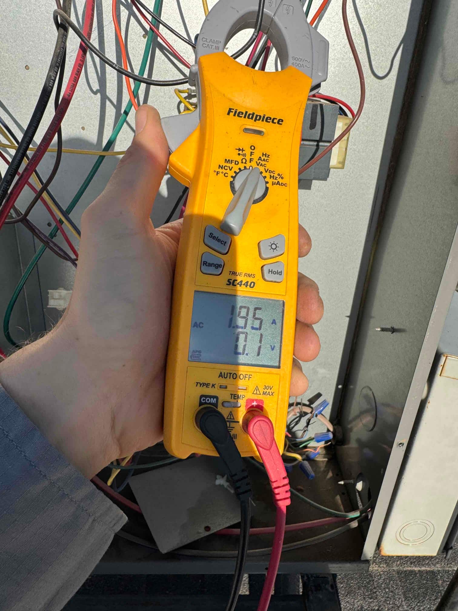 Clamp meter set to AC volts measures 295.1 V across two conductors inside the outdoor condenser control compartment.