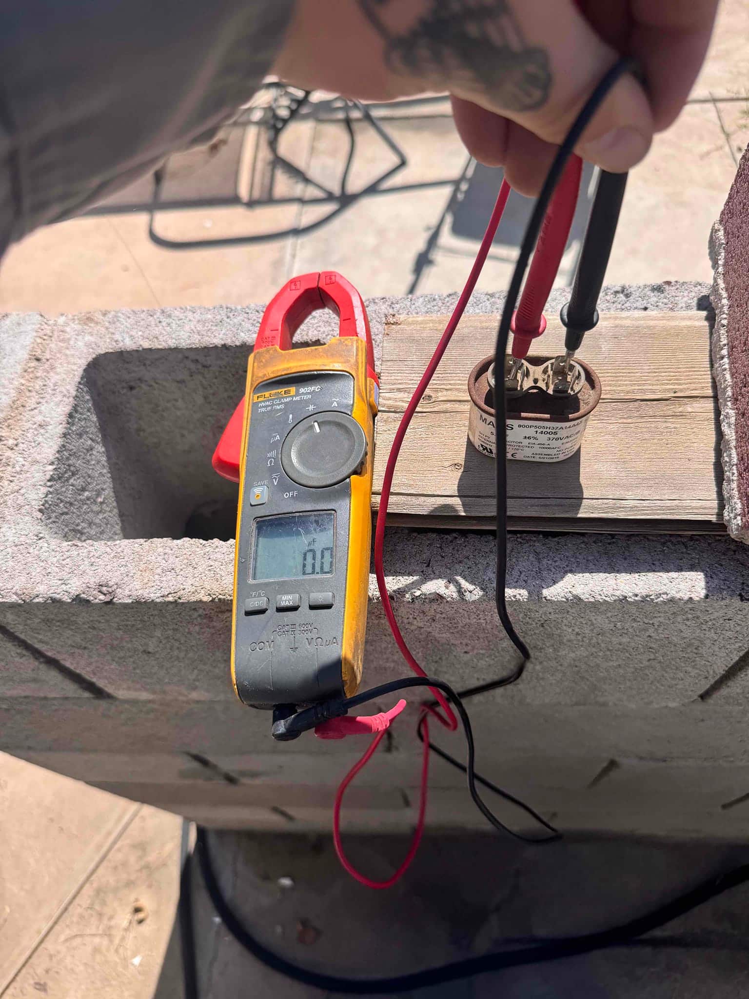 Clamp meter set to volts shows 0 V while probing a small run capacitor on a masonry block.