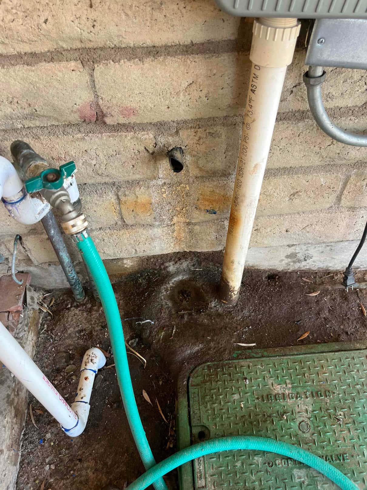 Condensate drain outlet at exterior wall with nearby PVC piping and garden hose, showing moisture staining below the discharge hole.