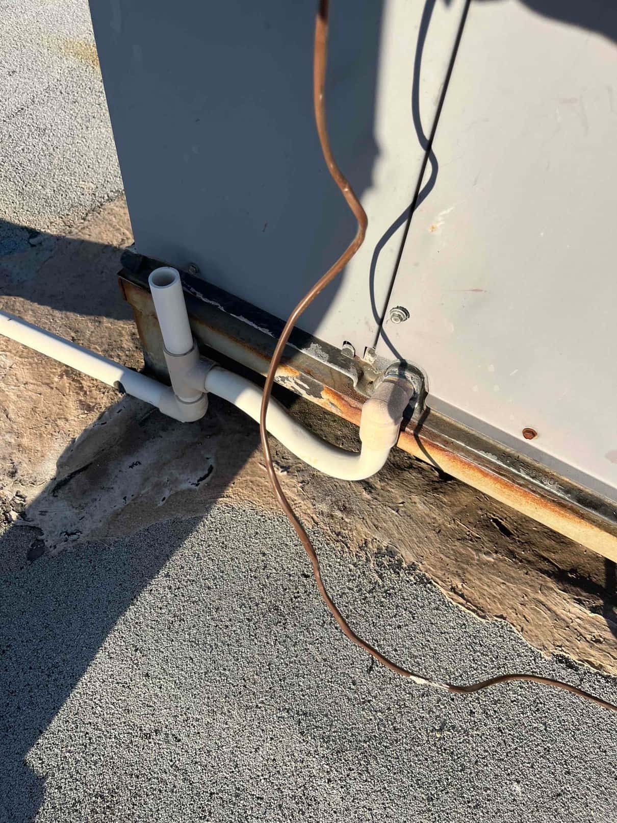Condensate drain piping and cleanout next to rooftop air handler cabinet, showing PVC connections and rusted base rail at the unit corner.