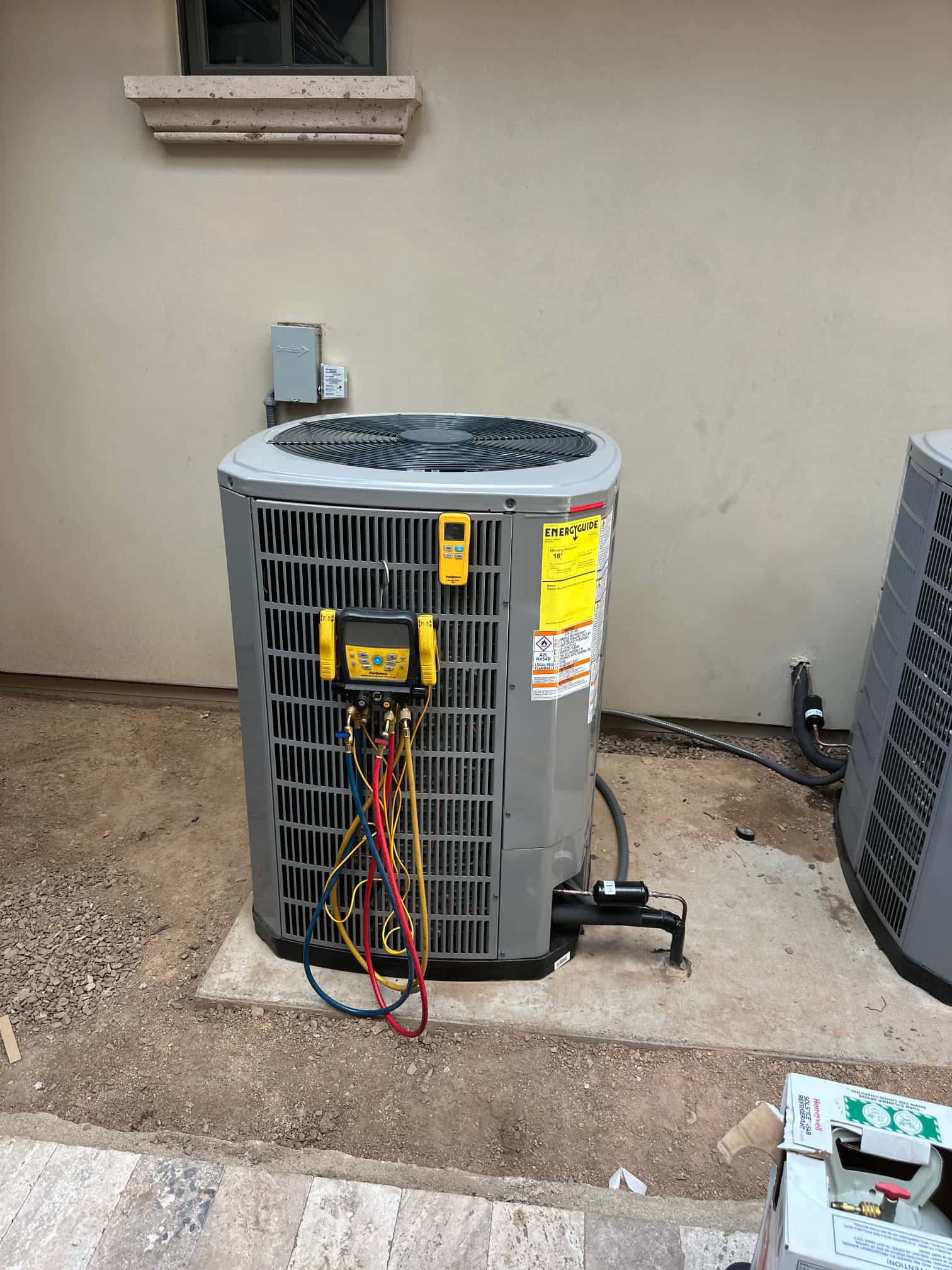 New outdoor condenser set on a level pad with manifold hoses connected and electrical disconnect mounted on the wall nearby.