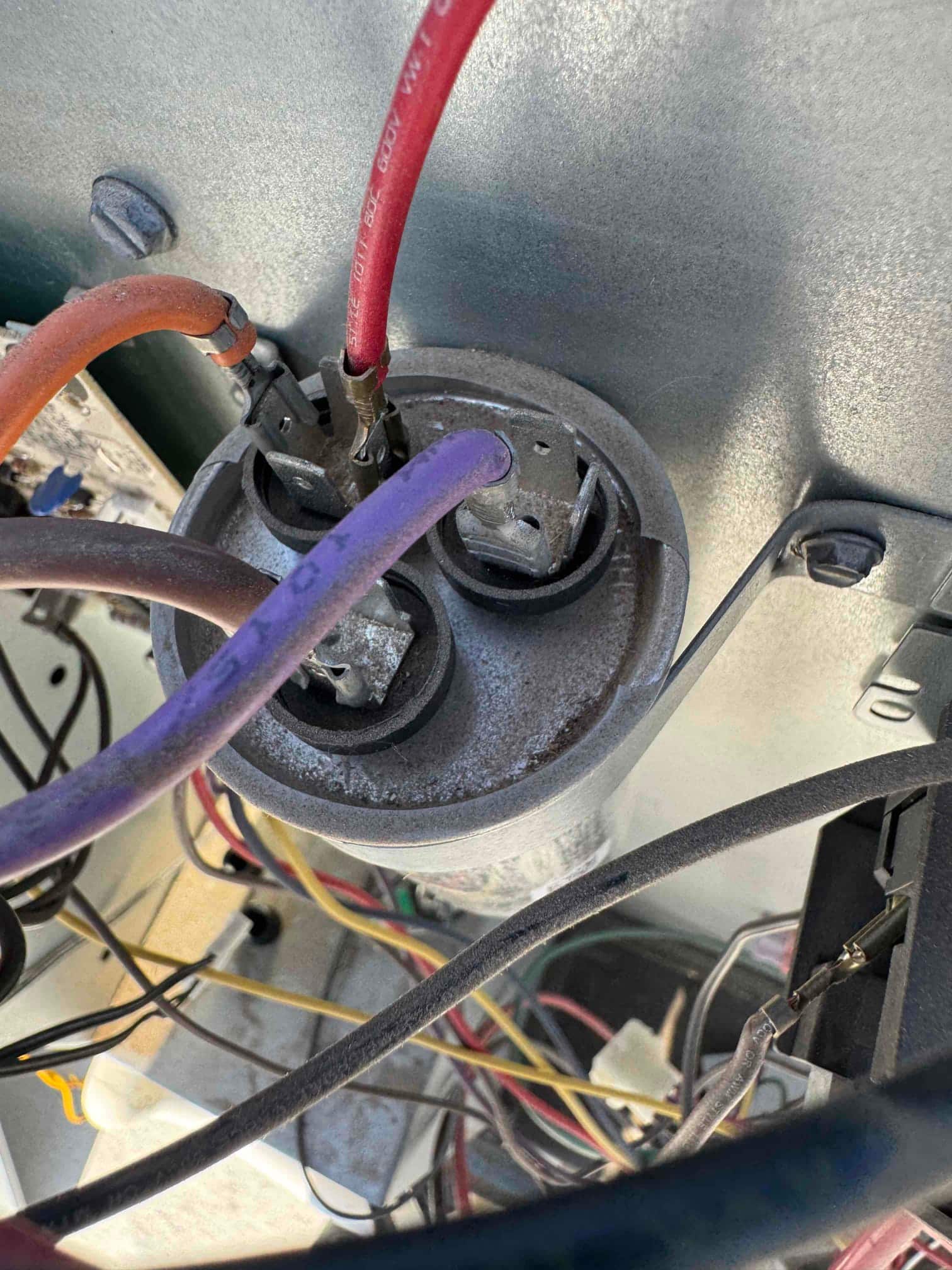 Outdoor condenser compartment showing dust-covered dual-run capacitor with connected red, orange, and purple wires on the terminals.