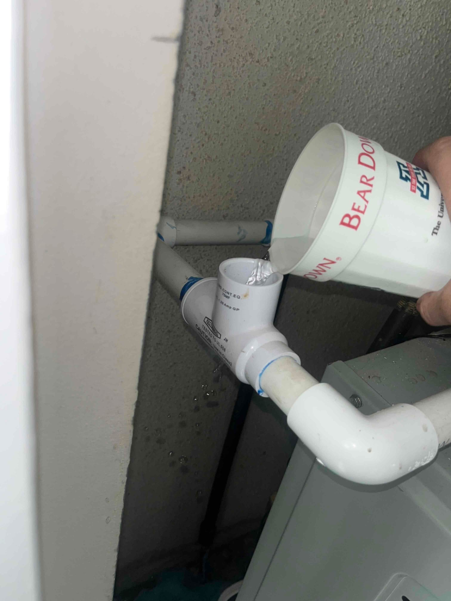 Technician pours water into a PVC condensate drain cleanout to verify flow through the repaired drain line.