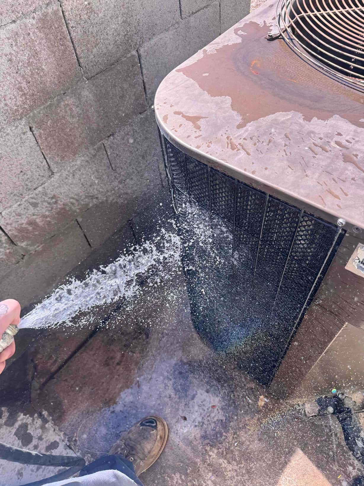 Technician rinses debris from an outdoor condenser coil with a hose, showing water spray across the coil fins.