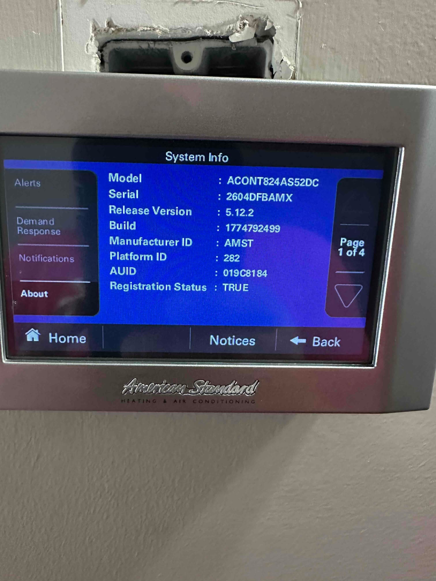 Wall-mounted touchscreen thermostat displays system information menu after installation, with housing not yet fully seated into the wall opening.