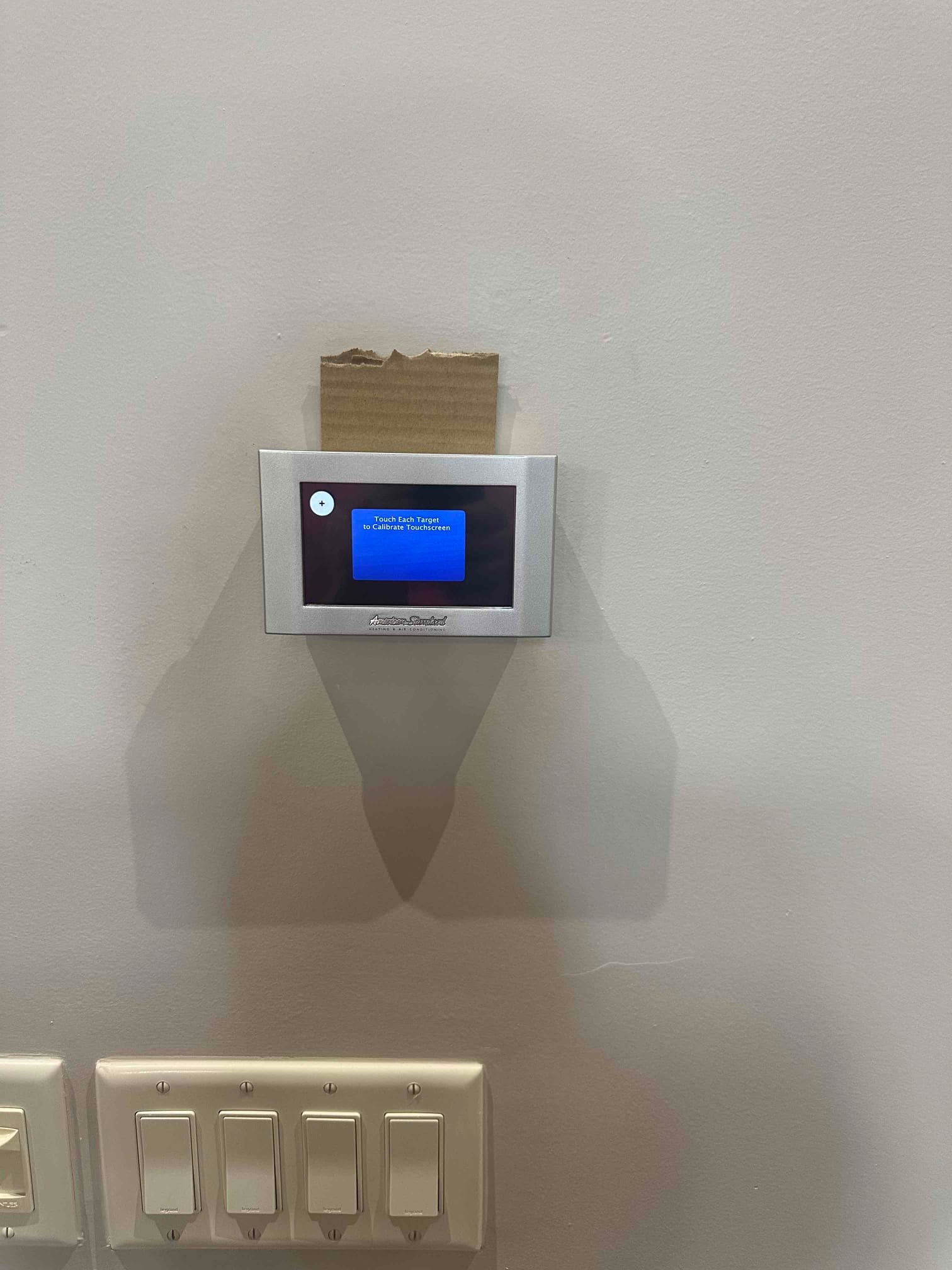 Wall-mounted touchscreen thermostat installed and powered on, displaying a calibration prompt above nearby light switches.