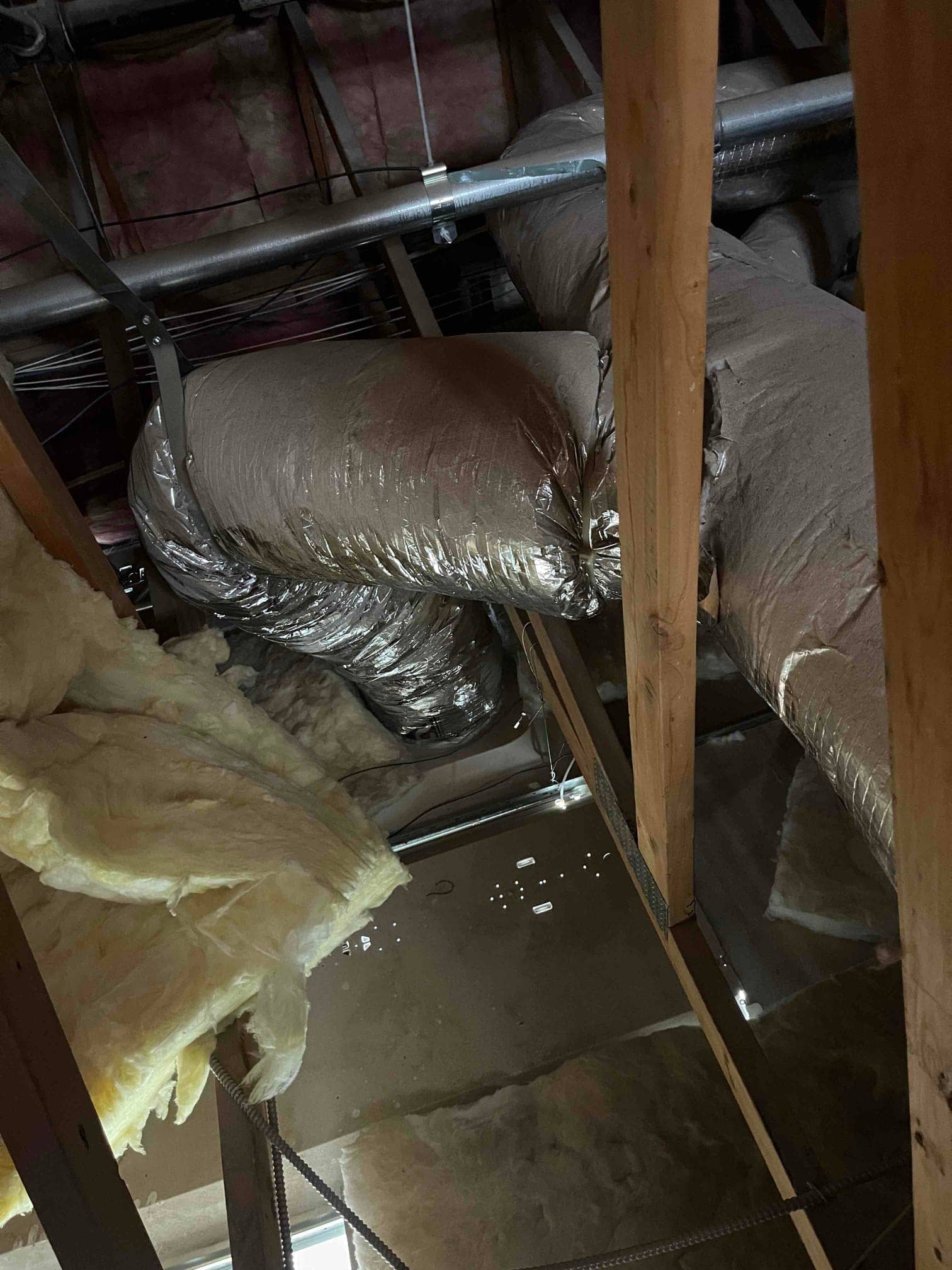 Attic air duct with foil insulation runs above a wet area on the ceiling below, showing moisture accumulation.