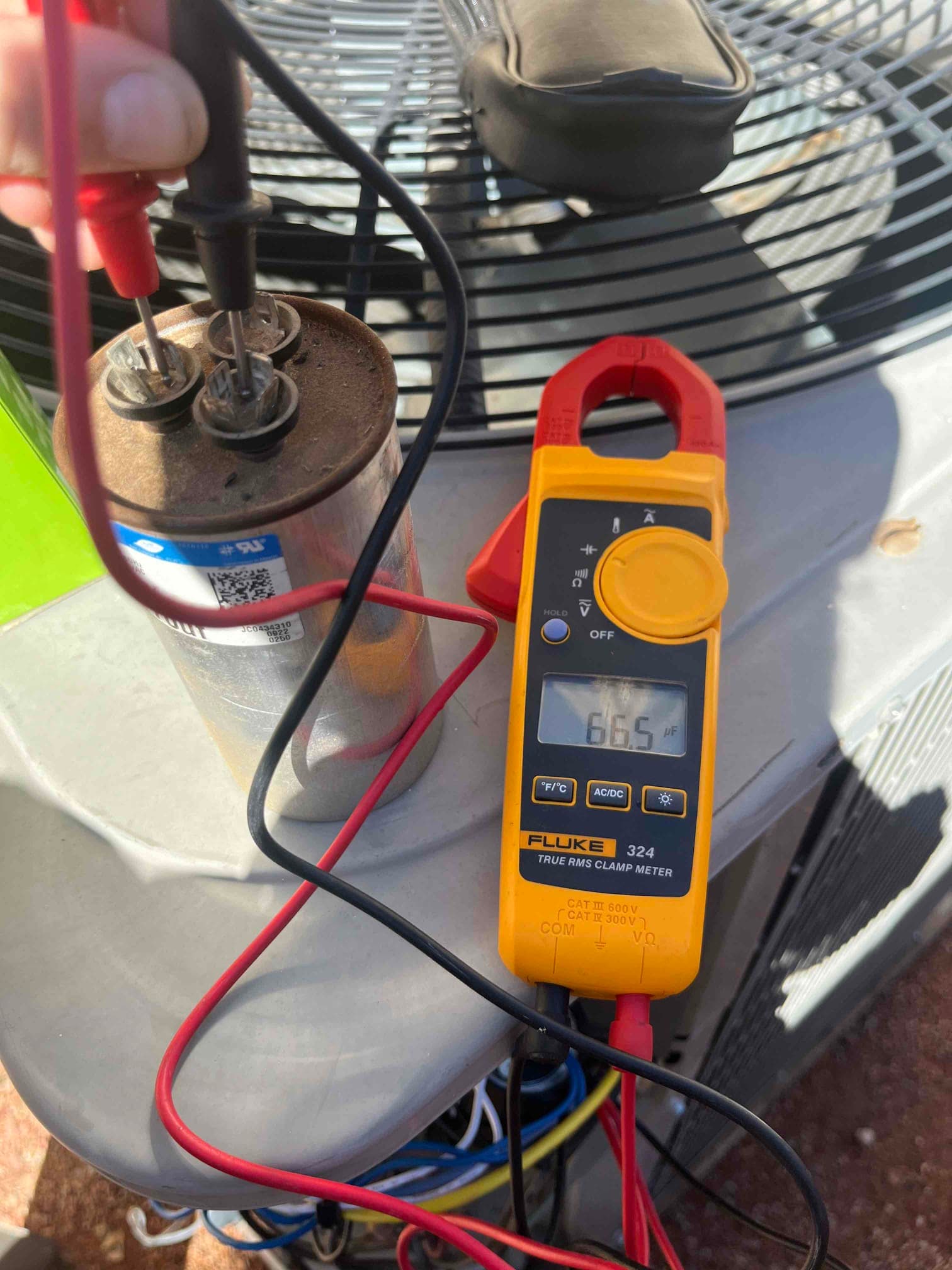 Clamp meter measures 66.5 μF on the dual-run capacitor using test leads, with the outdoor condenser unit in the background.