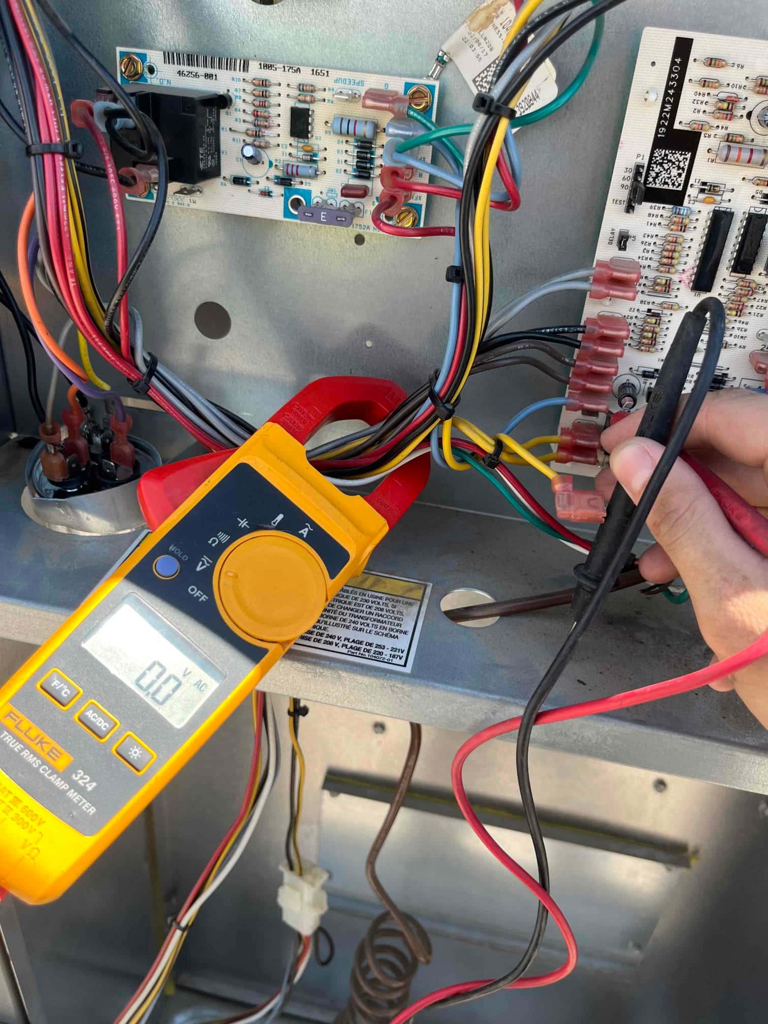 Clamp meter measures voltage at a control board terminal inside the condenser panel, displaying 0.0 V on the screen.